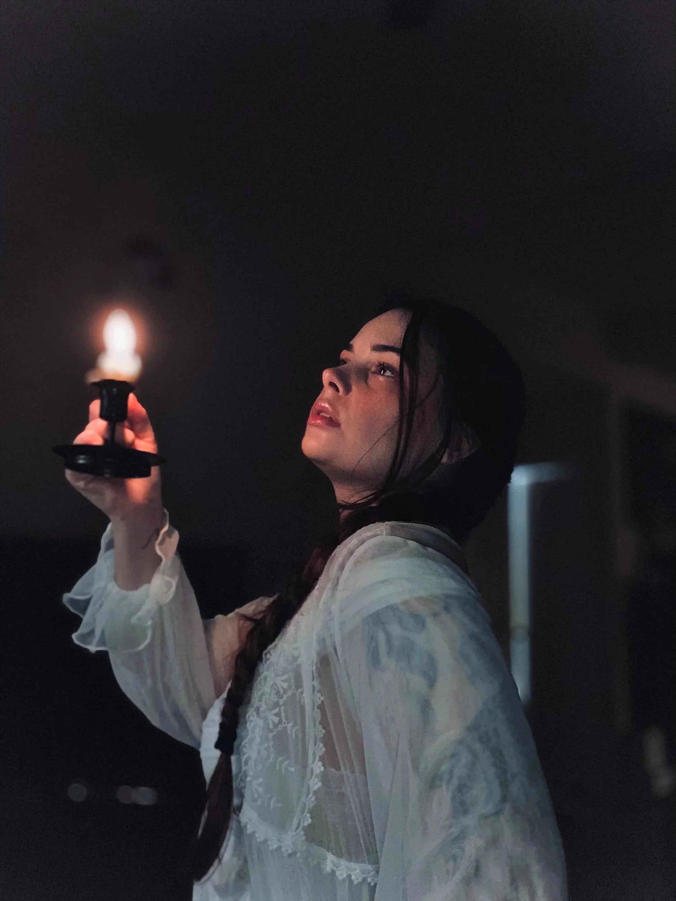 A woman with dark hair in a braid holds a lit candle in a dark room, looking upward with a contemplative expression, wearing a white lace blouse.