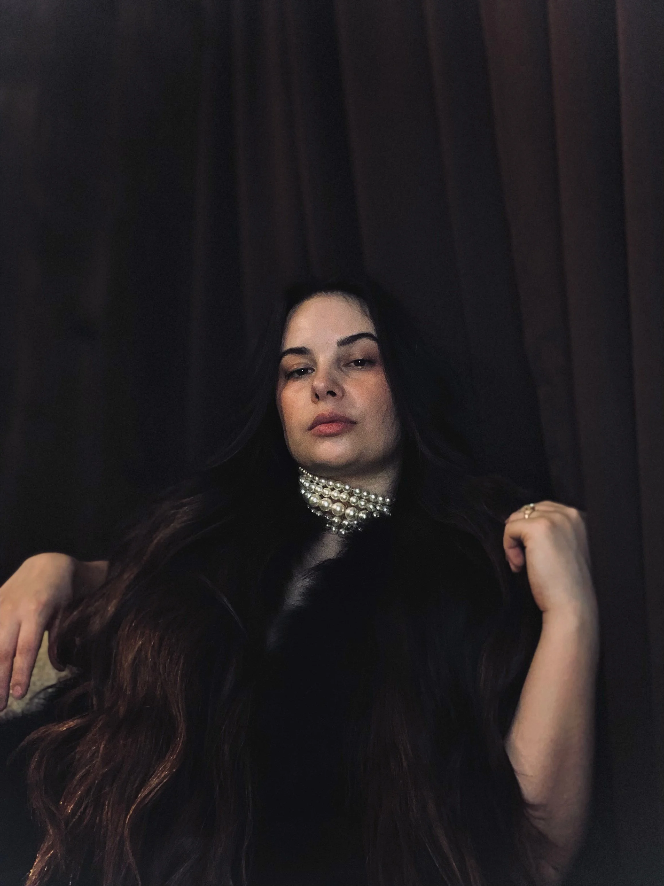 A woman with long dark hair wearing a pearl choker necklace and a black outfit, posing against a dark wooden background.