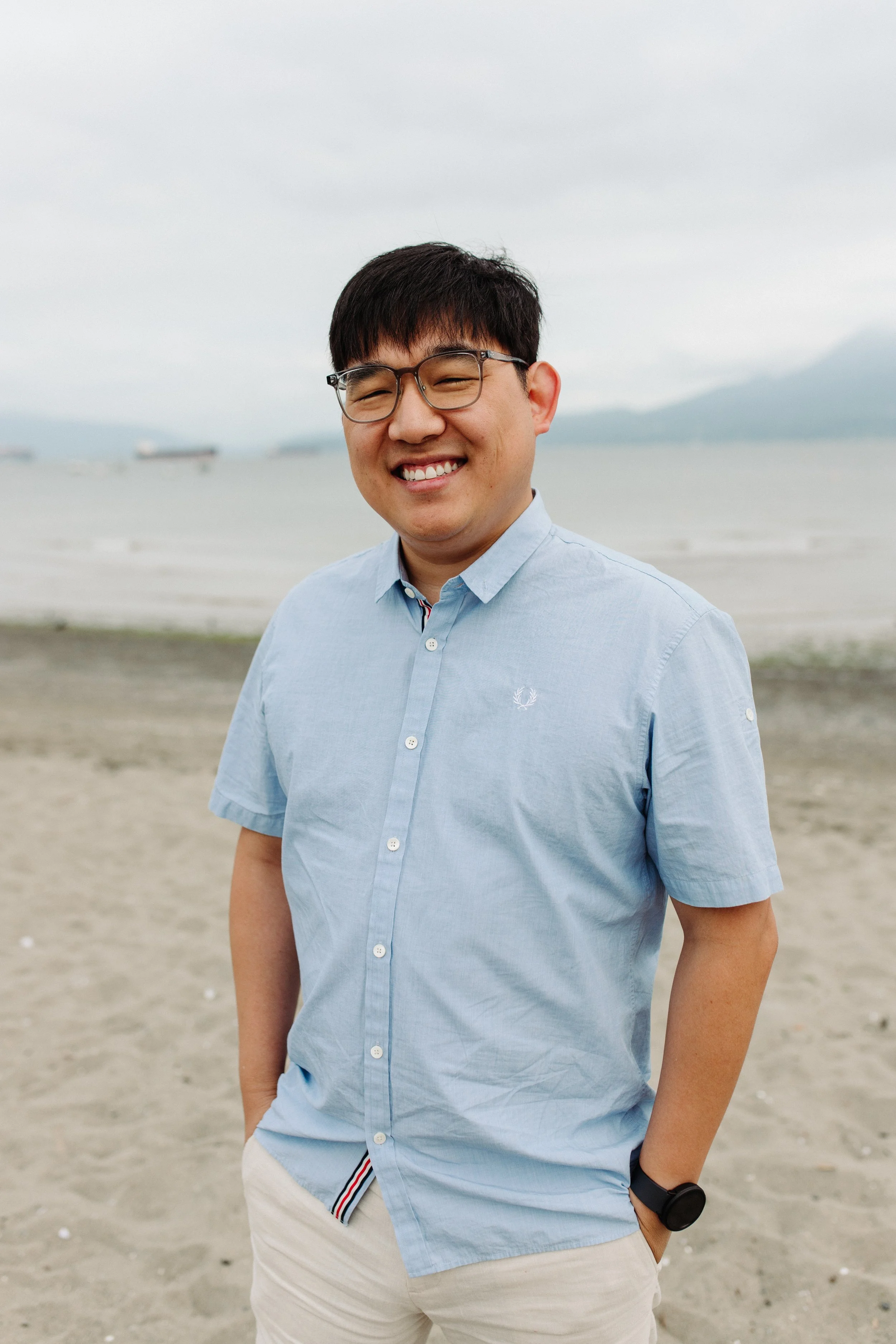 A smiling man in a light blue short-sleeved shirt and beige shorts standing on a beach with water and boats in the background.