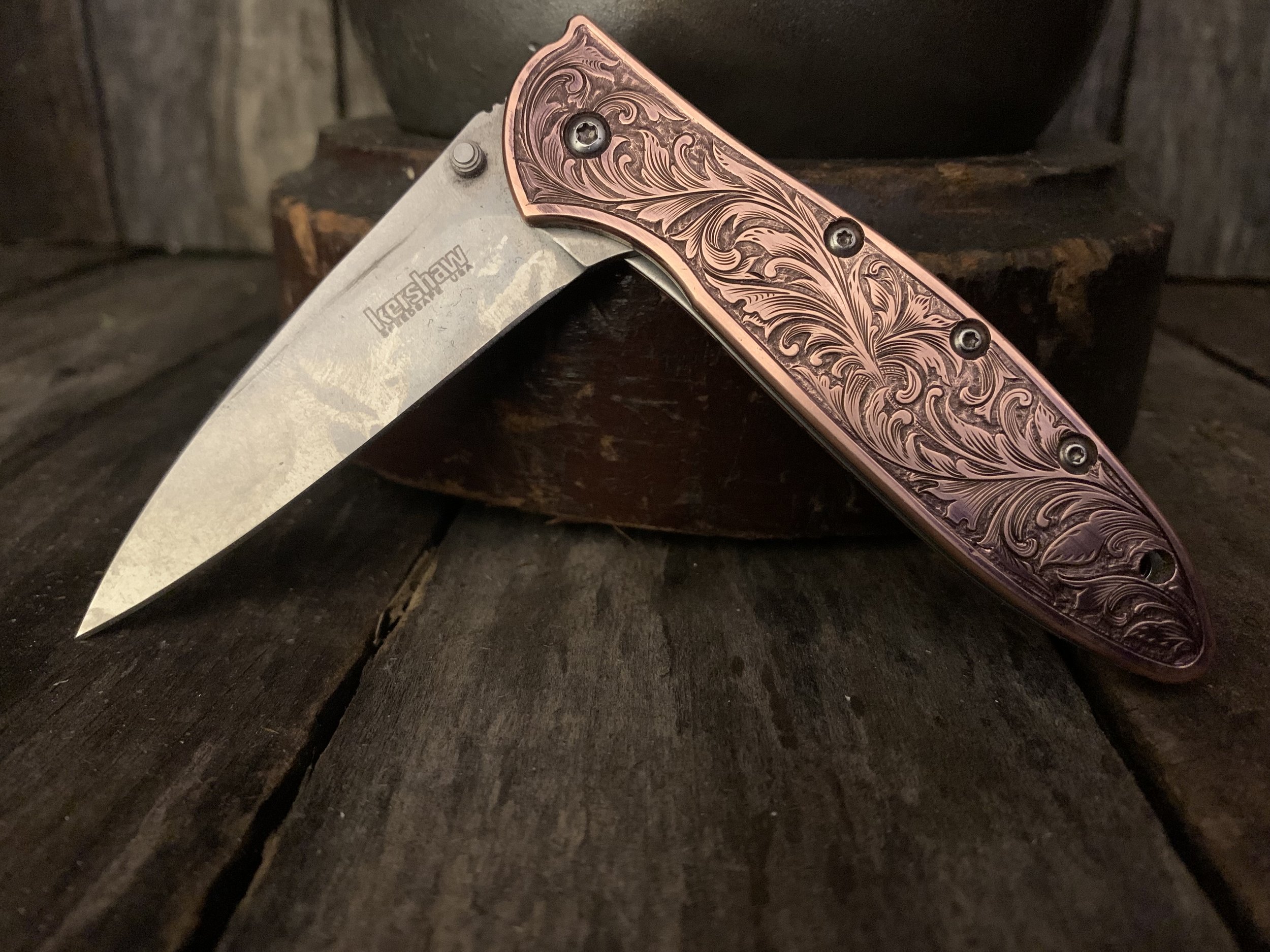 A folding pocket knife with a decorative, engraved handle and a partly open blade on a rustic wooden surface.