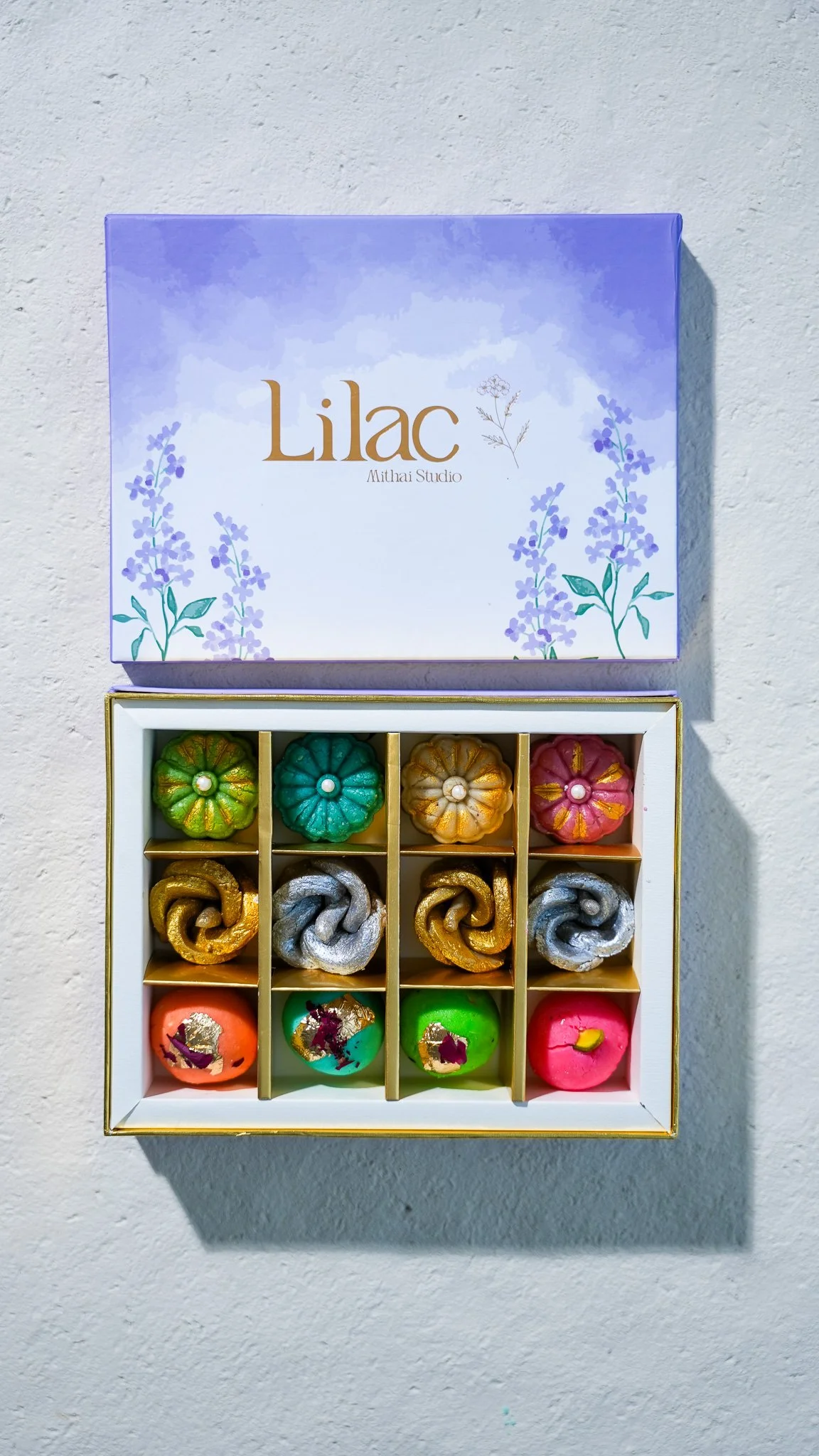 Colorful decorative desserts in a white display box with 12 compartments, filled with various brightly colored confections resembling fruits, flowers, and abstract shapes.