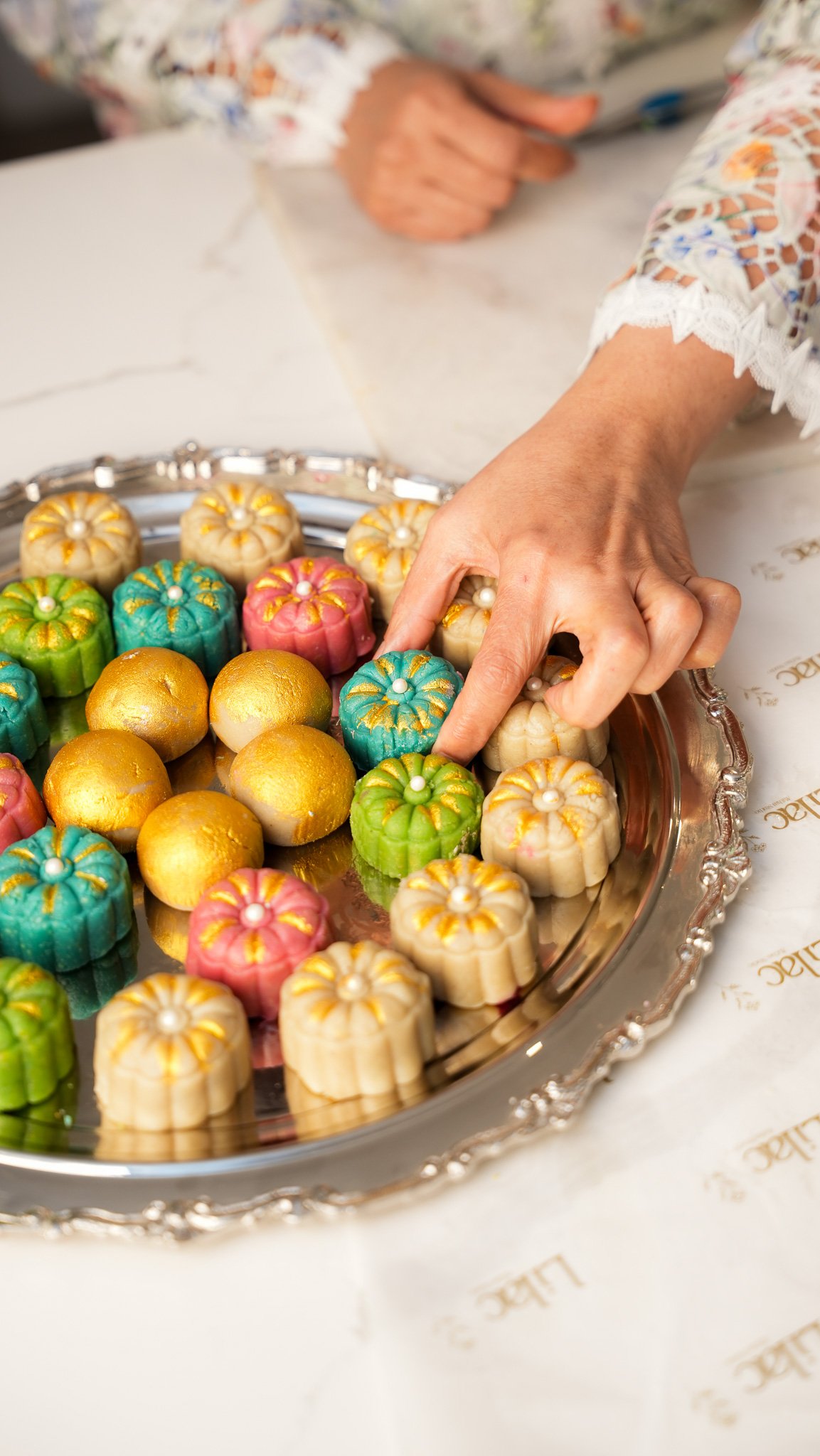 A hand reaching to pick up colorful flower-shaped and gold-colored desserts on a silver platter.