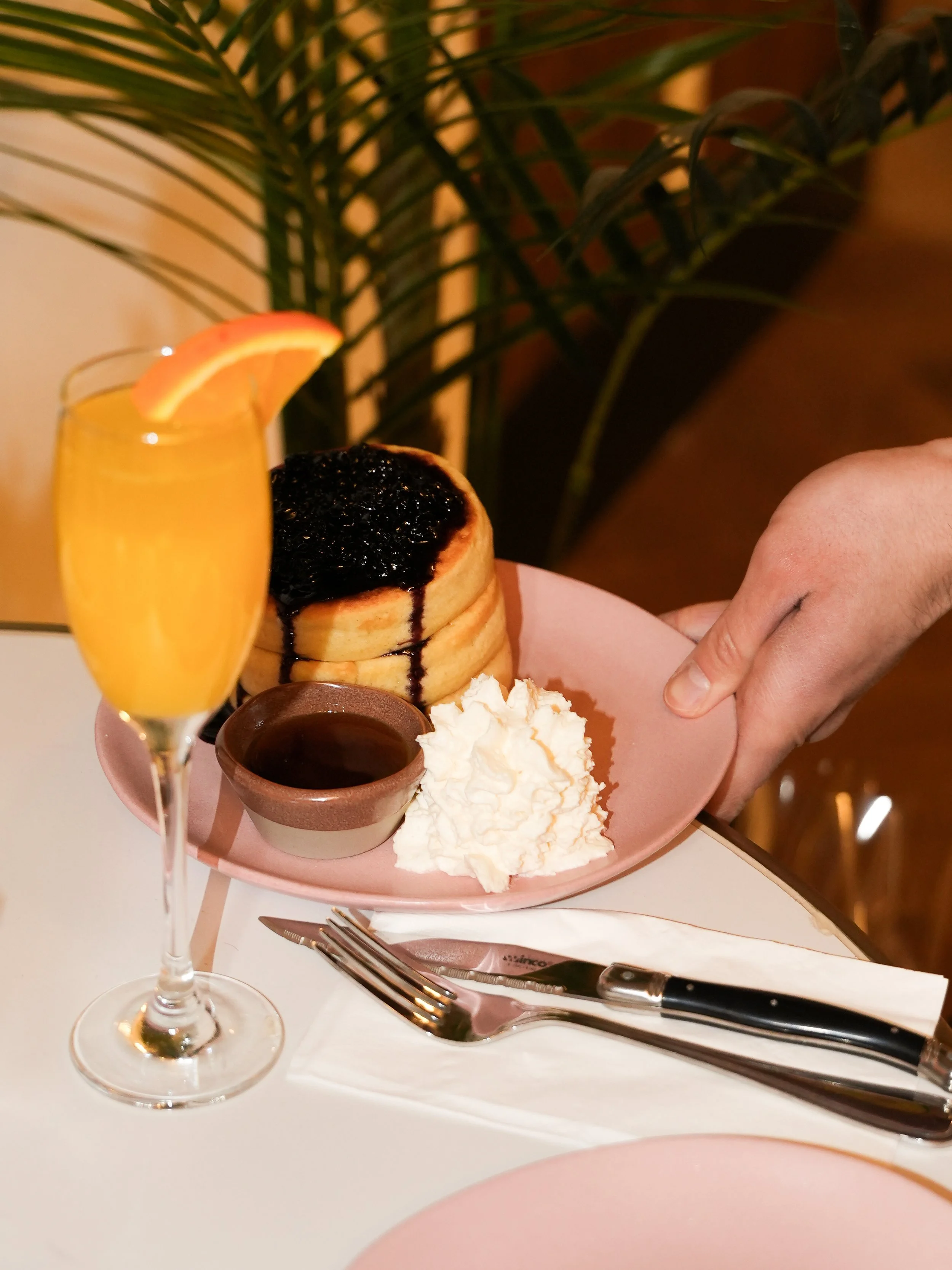 A plate of fluffy pancakes topped with blueberry compote, served with whipped cream, chocolate sauce in a small bowl, an orange slice in a glass of orange juice, and utensils on a white table with a pink plate in the background, against a backdrop of