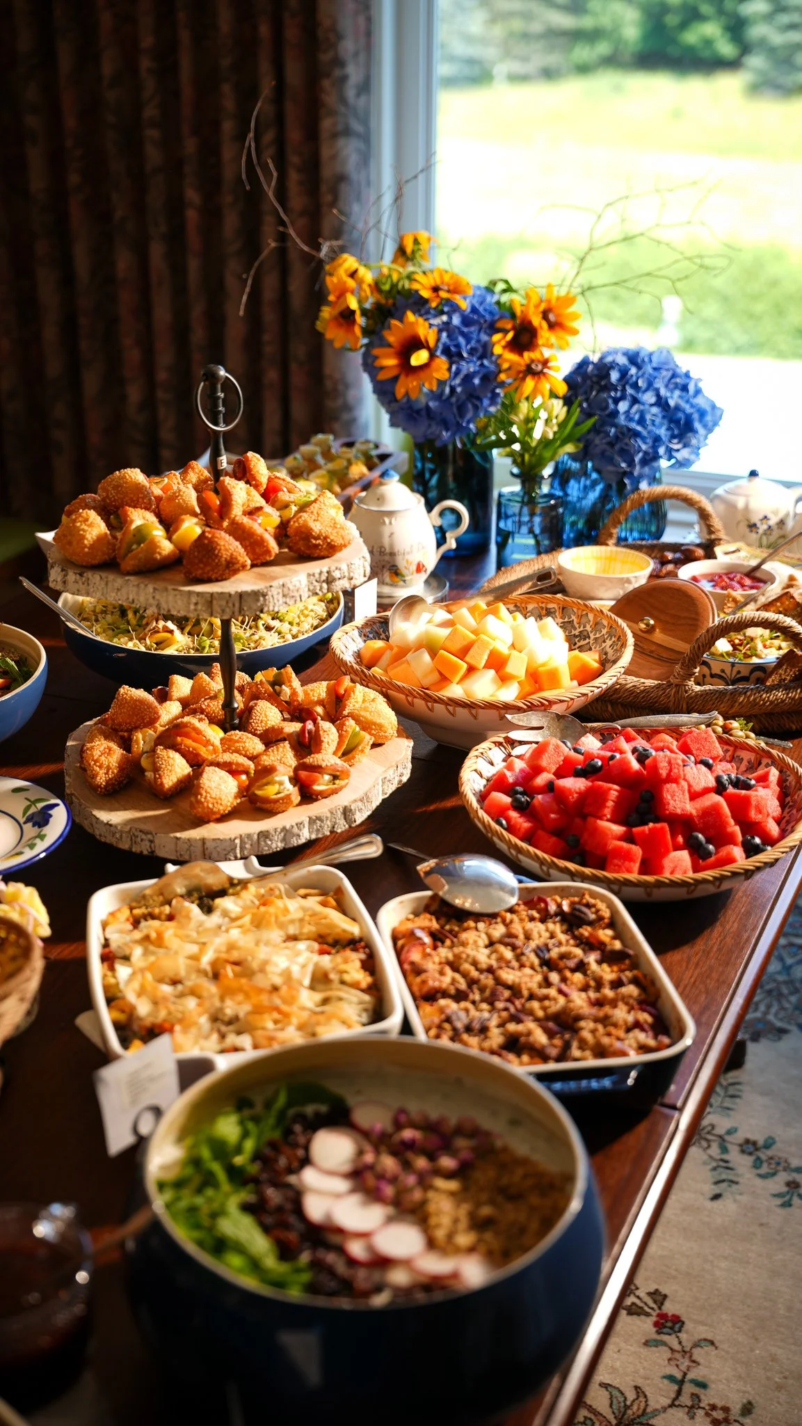 A table filled with various dishes including fruit salads, baked goods, and casserole dishes, with a vase of vibrant flowers in the background near a window.