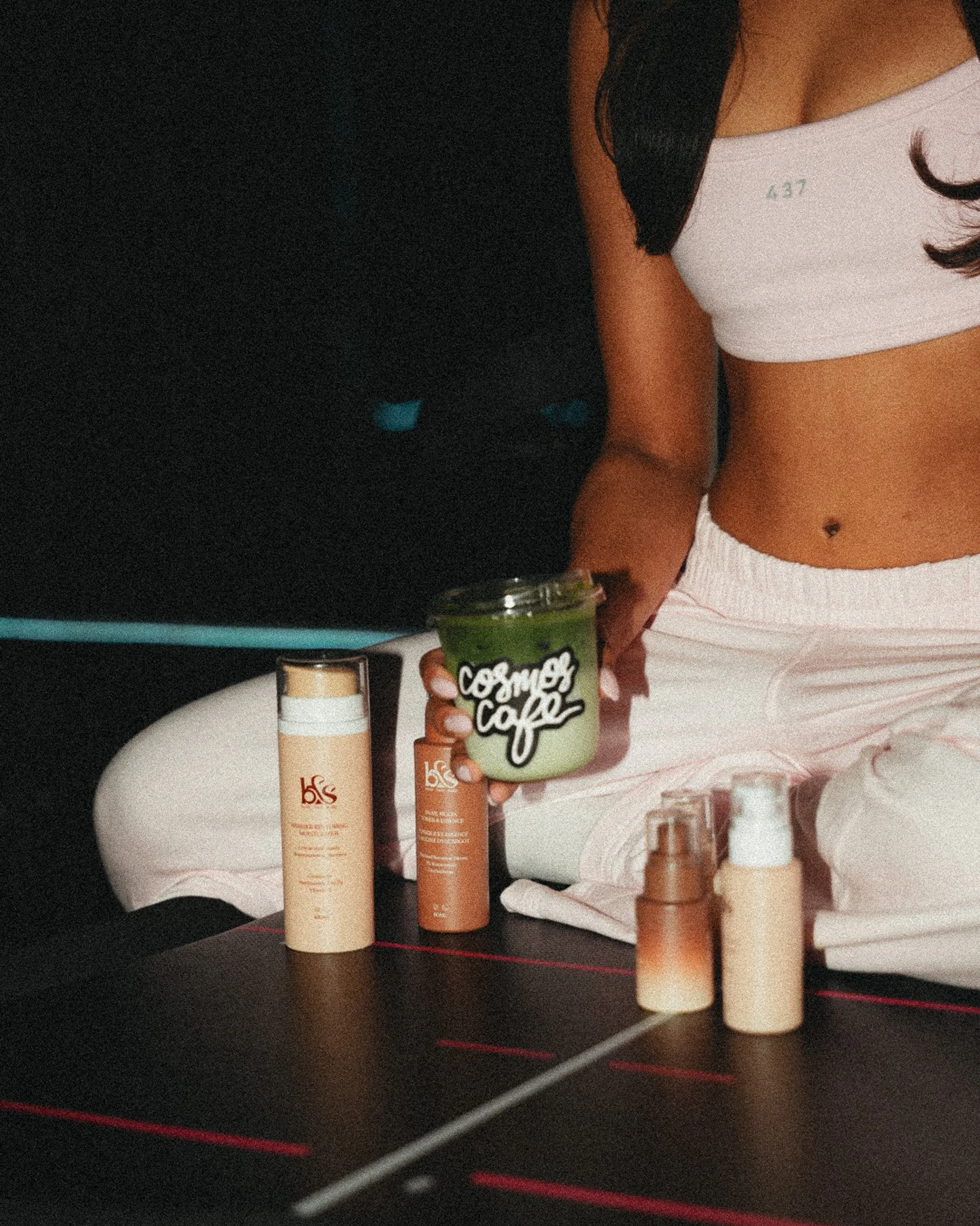 A woman standing in front of a black countertop holding a green beverage with a straw in a clear cup with the words 'Comfort Coffee' on it. Various skincare or cosmetic products are arranged on the countertop, including bottles and jars.