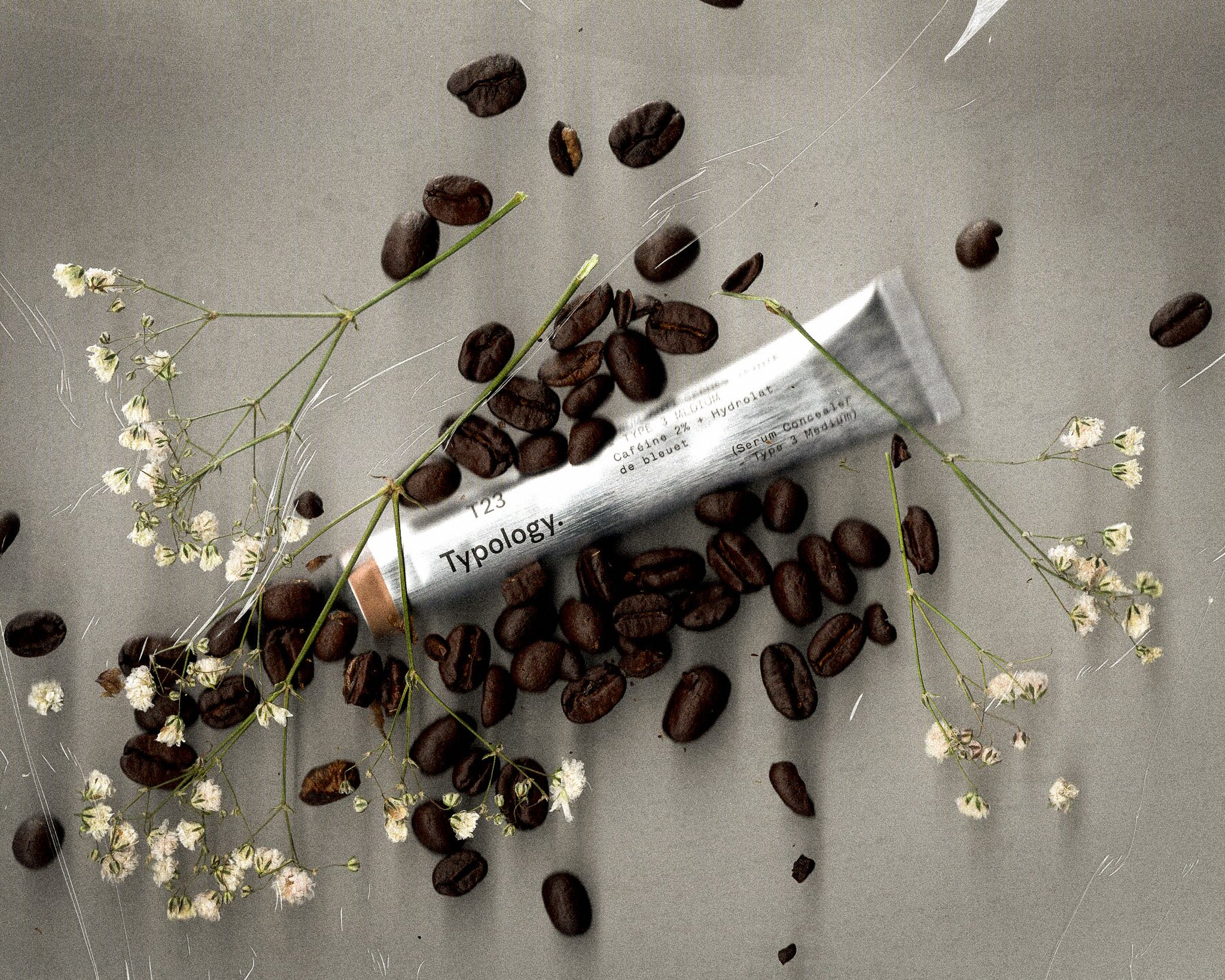 Coffee beans scattered around a tube labeled 'Typology' on a gray surface, with small white flowers entwined among the beans.