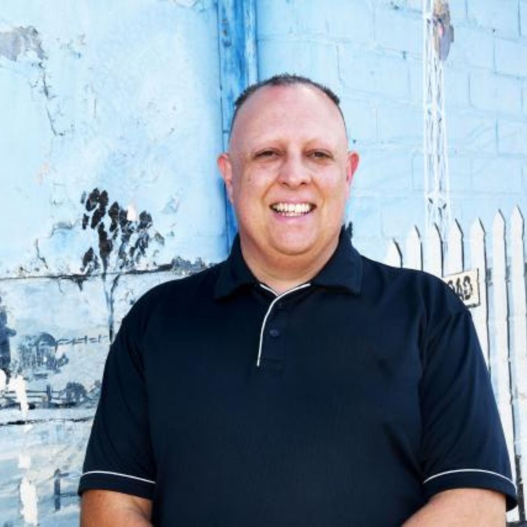 A smiling man standing in front of a weathered wall with paint peeling, wearing a black polo shirt.