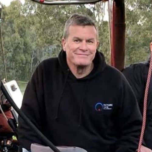A middle-aged man with gray hair smiling, wearing a black hoodie with a logo on the chest, outdoors with trees in the background.