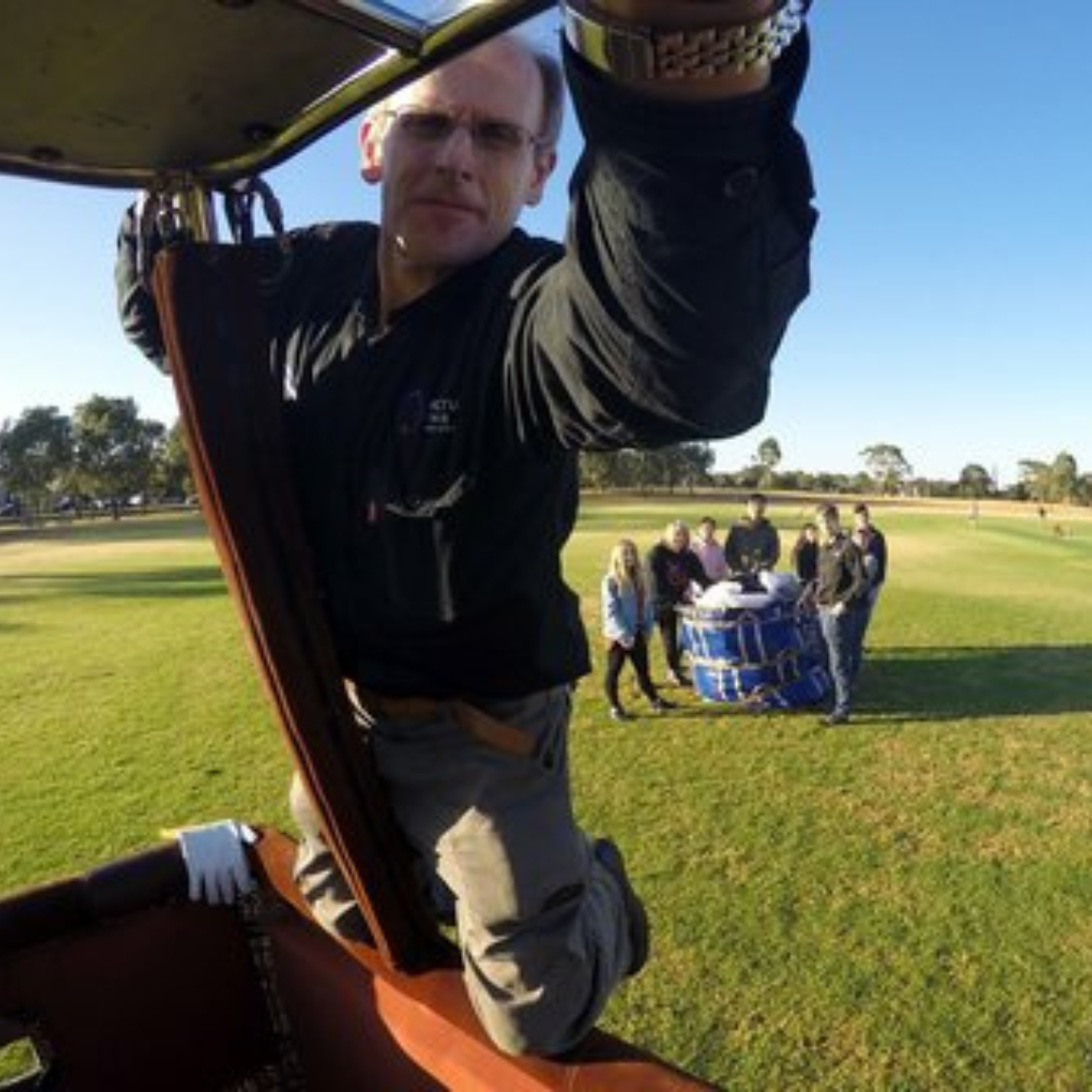 A man taking a selfie while kneeling inside a hot air balloon basket, with a group of six people standing in the background on a grassy field near the balloon.
