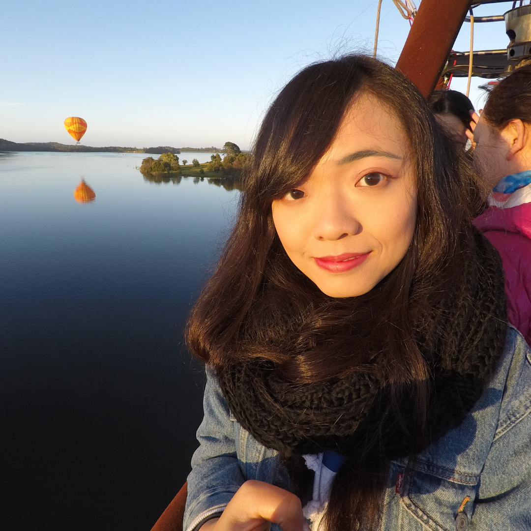 A woman with dark hair smiling as she takes a selfie on a hot air balloon ride over a calm body of water with a hot air balloon in the distance, during sunset.