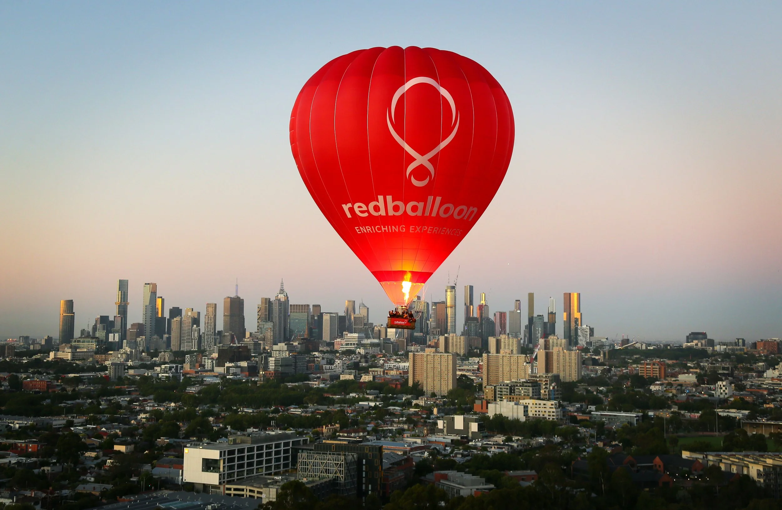 Red hot air balloon with the logo and text 'redballoon enriching experiences' floating above a city skyline during sunset.