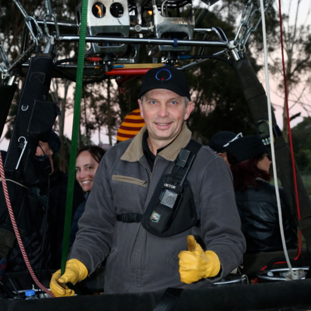 A man smiling and giving a thumbs-up in a hot air balloon basket, with three women inside. The basket contains equipment and the sky is visible in the background.