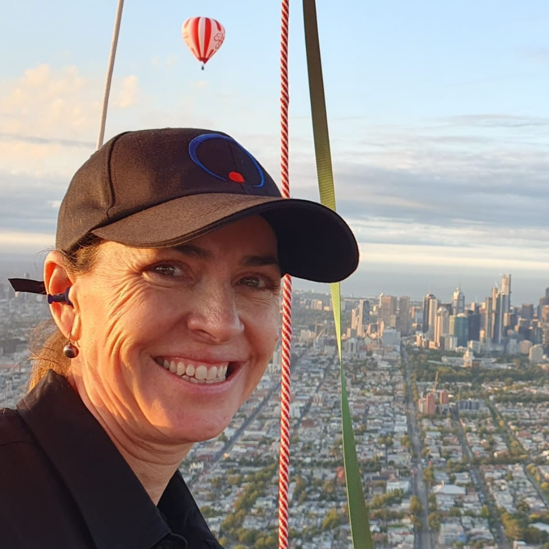 A woman smiling in a hot air balloon with a city skyline in the background and a red and white striped hot air balloon in the sky.