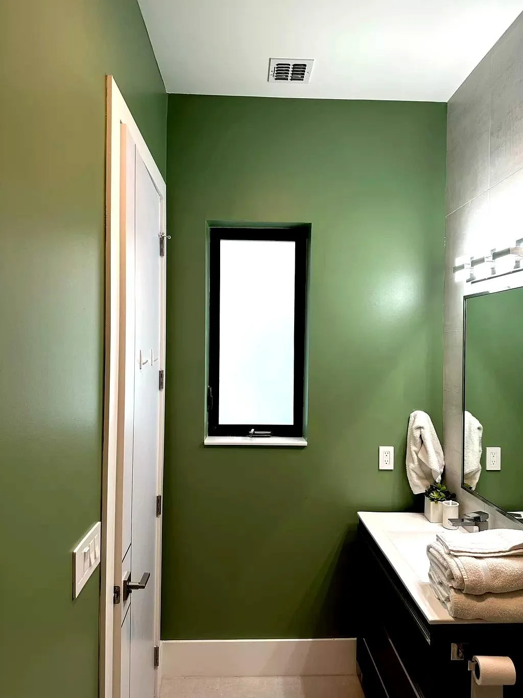 Bathroom with green walls, a small window, a mirror, a dark vanity with folded towels, and a towel hanging on the wall.