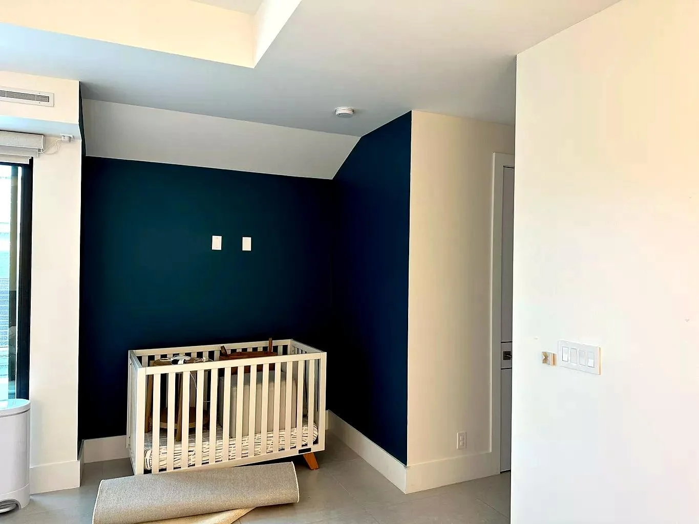 Empty nursery with a white crib against a dark blue accent wall and a beige rug on the floor.