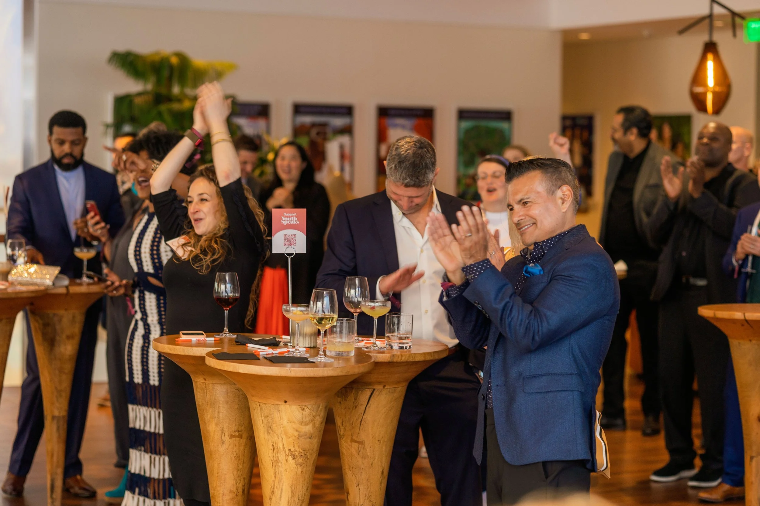 A group of people at a social event, some are clapping, others are taking photos or raising their hands in celebration, with drinks on tables in a warmly lit reception area.