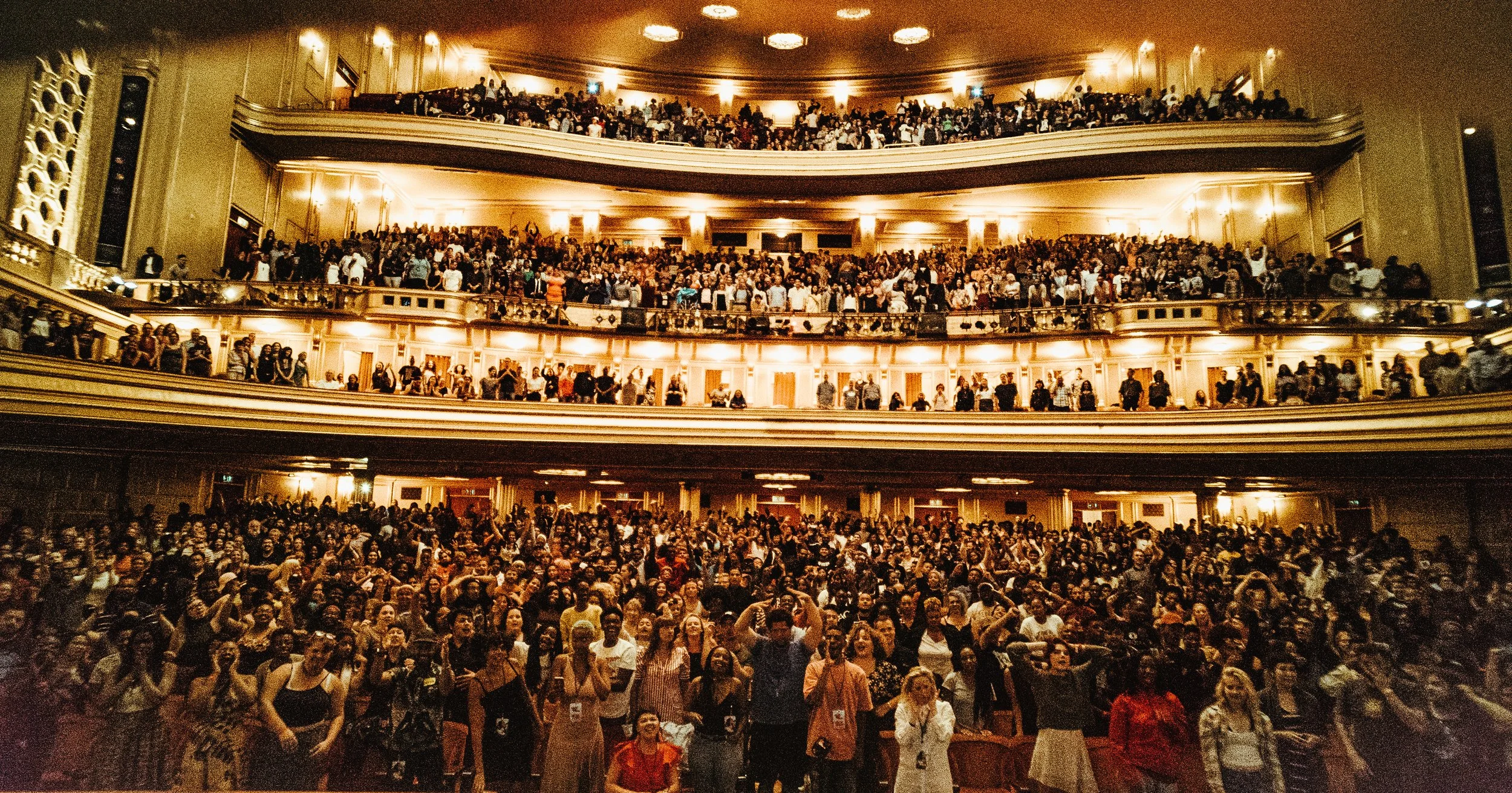 Large theater filled with a crowd of people, some seated and some standing, with multiple balconies and warm lighting.