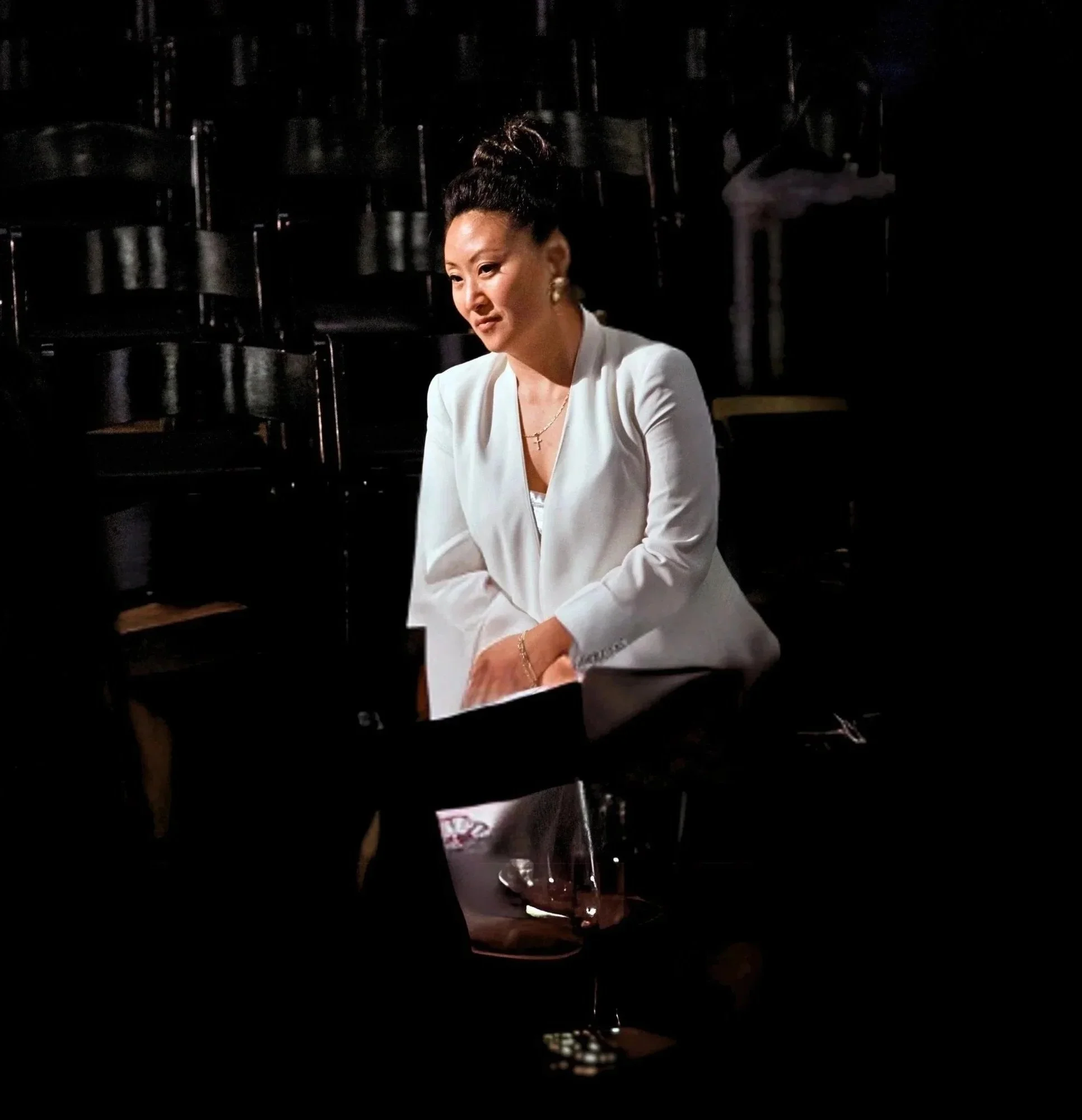 A woman with dark hair styled in a bun, wearing a white blazer, earrings, and a necklace, listening in dark theater.