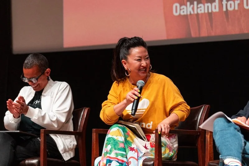 Three people seated on stage, engaging in a panel discussion. The woman in the center wearing a mustard yellow sweater with a colorful skirt is holding a microphone and laughing. The person on the left wearing glasses and a white jacket is clapping and smiling. The background features a screen with text partially visible, including the words "Oakland for."