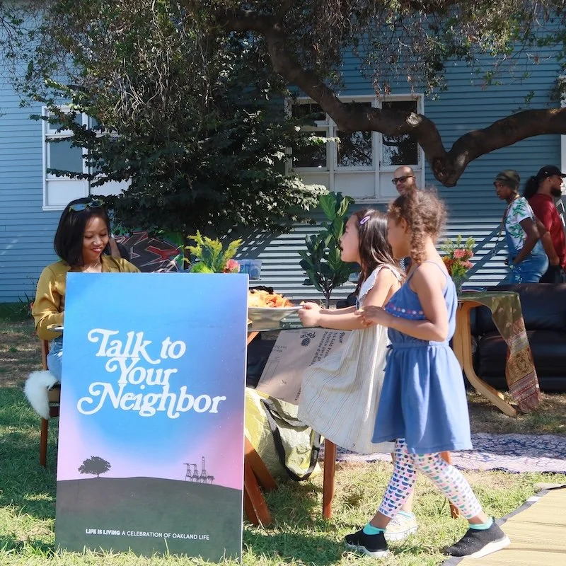 Three children talking to a woman at a neighborhood event, with a sign that says 'Talk to Your Neighbor,' outdoors with trees and houses in the background.