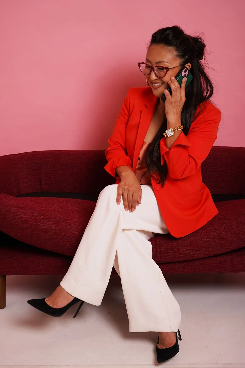 A woman with long dark hair, glasses, and wearing a red blazer and cream pants, sitting on a red couch against a pink wall, talking on a mobile phone.