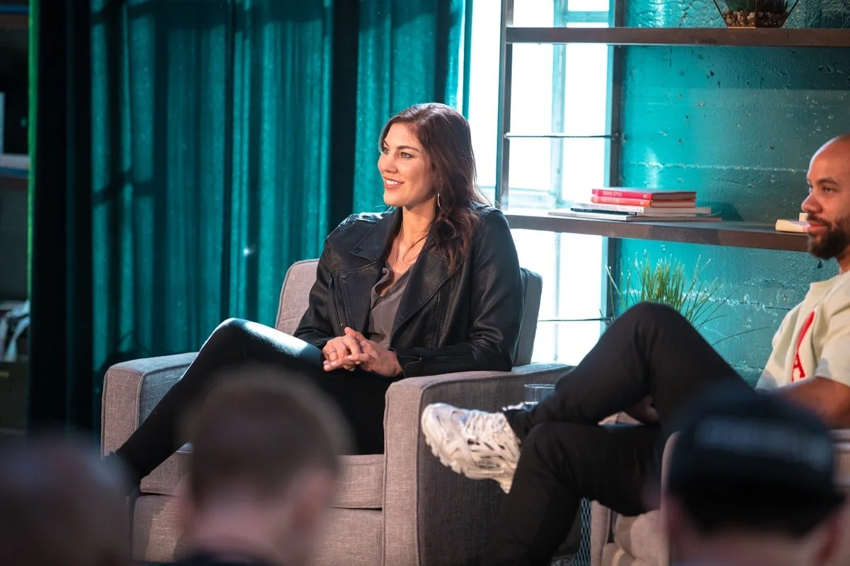 A woman in a leather jacket sitting on a gray armchair, smiling during a panel discussion or interview, with another person sitting nearby in a room with teal curtains and bookshelf.