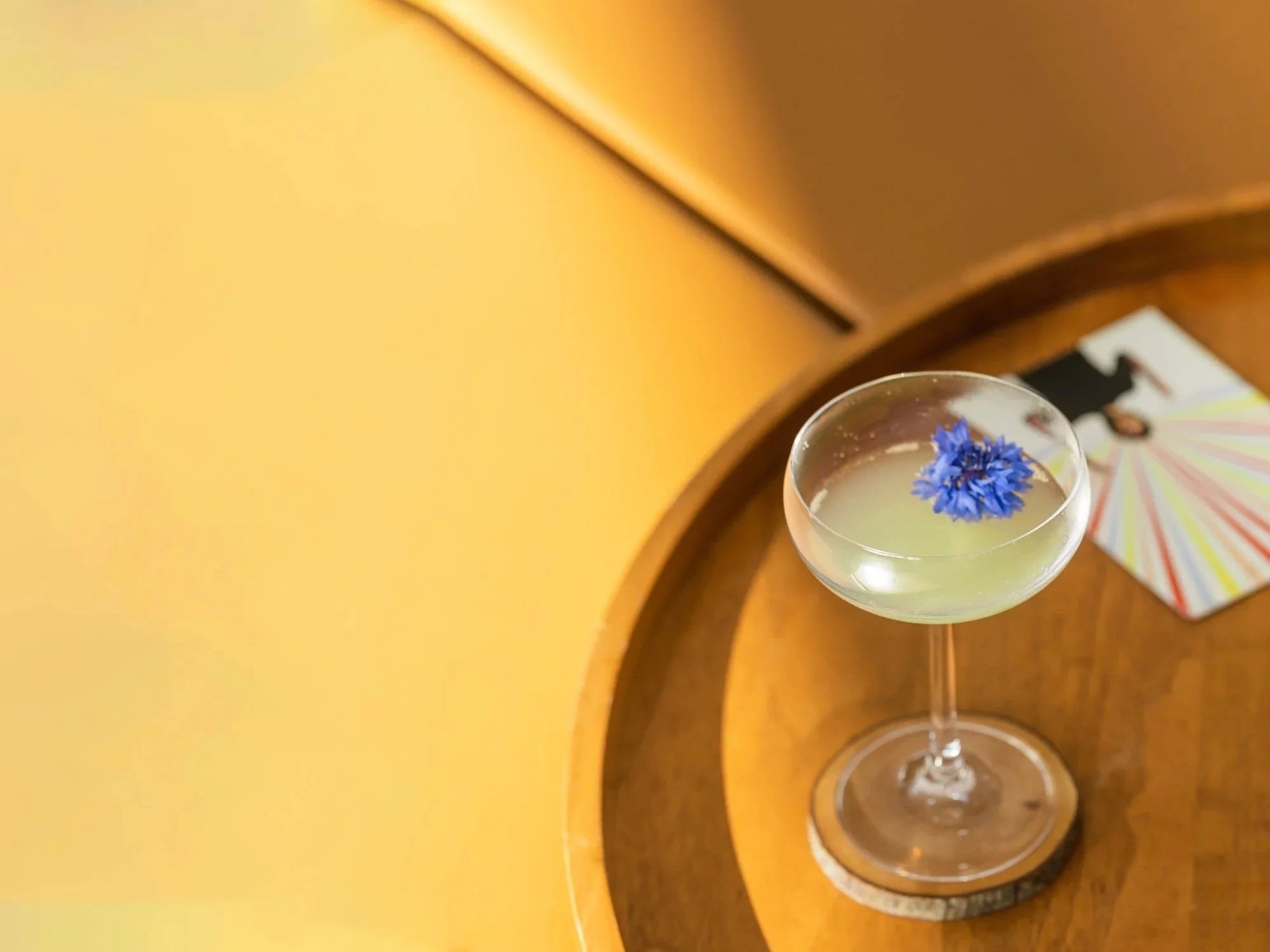 A cocktail glass with a yellow-green drink and a blue flower garnish on top, placed on a wooden tray with a colorful photo or postcard and a black light switch in the background.