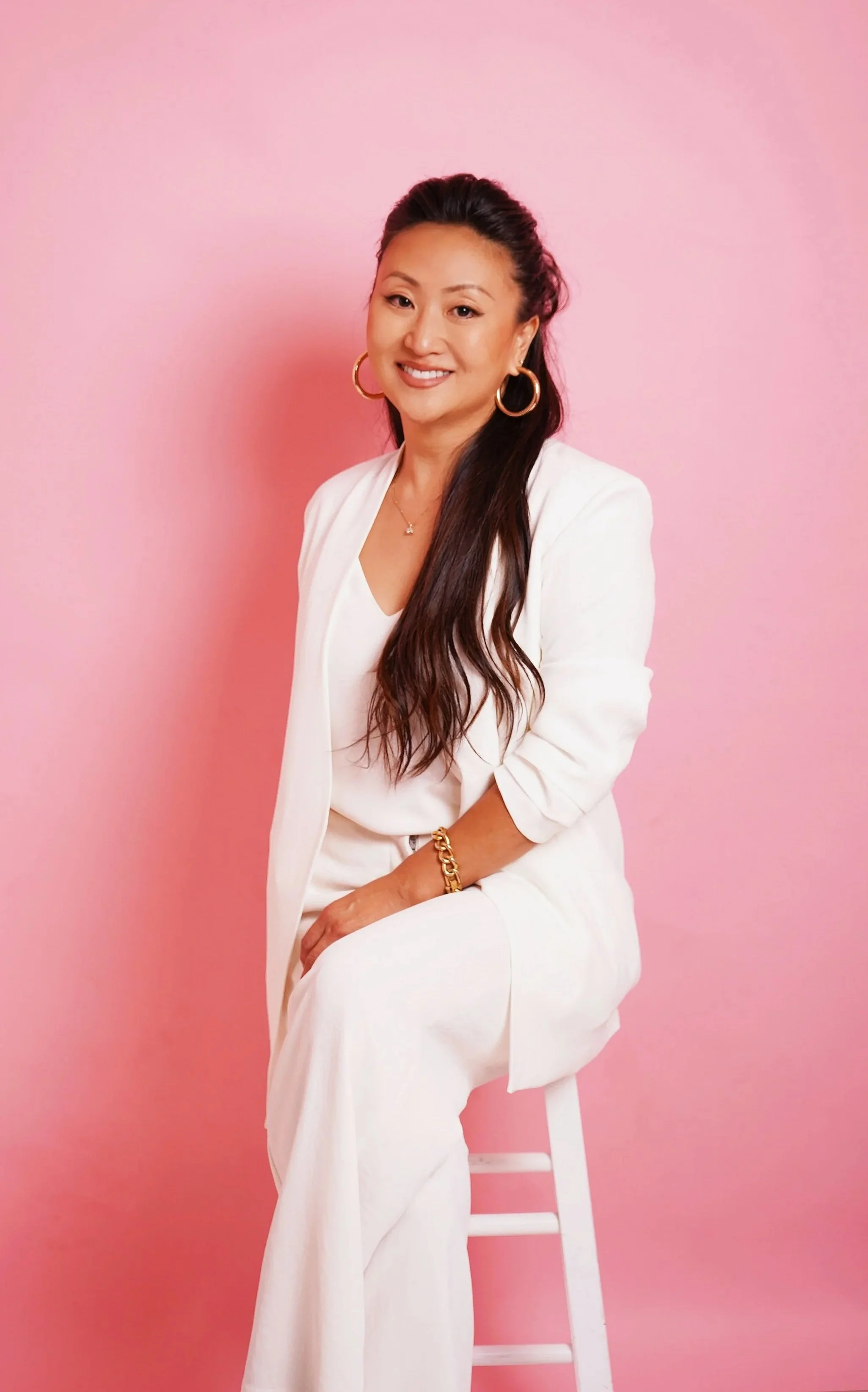 Woman in white suit sitting on a white stool against a pink background, smiling, with long dark hair, hoop earrings, and gold jewelry.