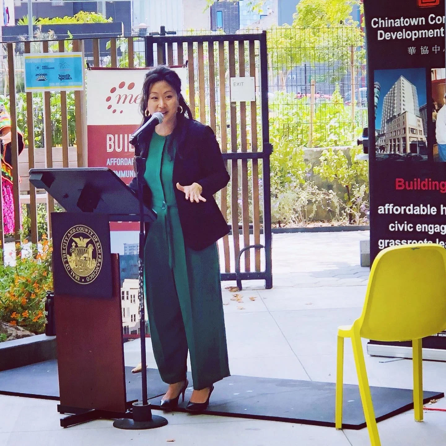 A woman speaking at a podium outdoors during a public event, with a woman in traditional attire visible in the background, banners, and a bright yellow chair nearby.