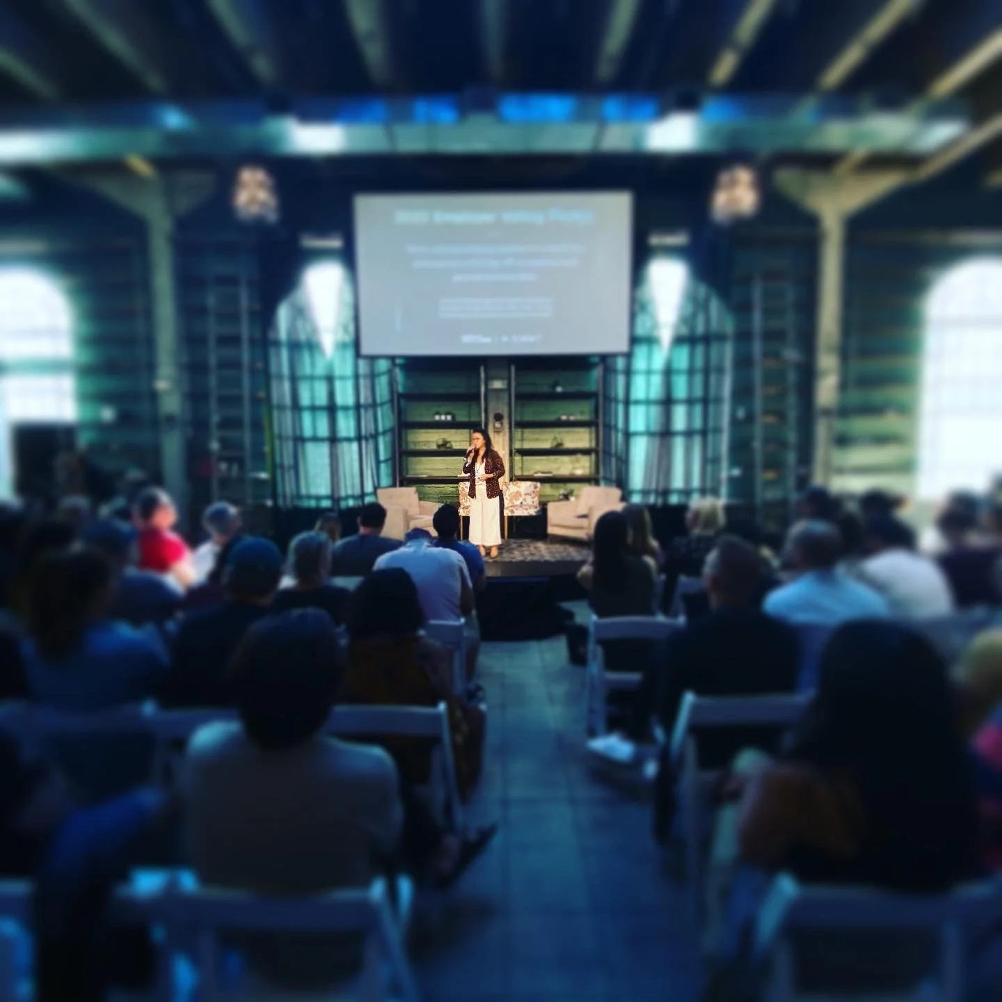 A woman giving a presentation or speech on stage in front of an audience inside a glass-walled building.