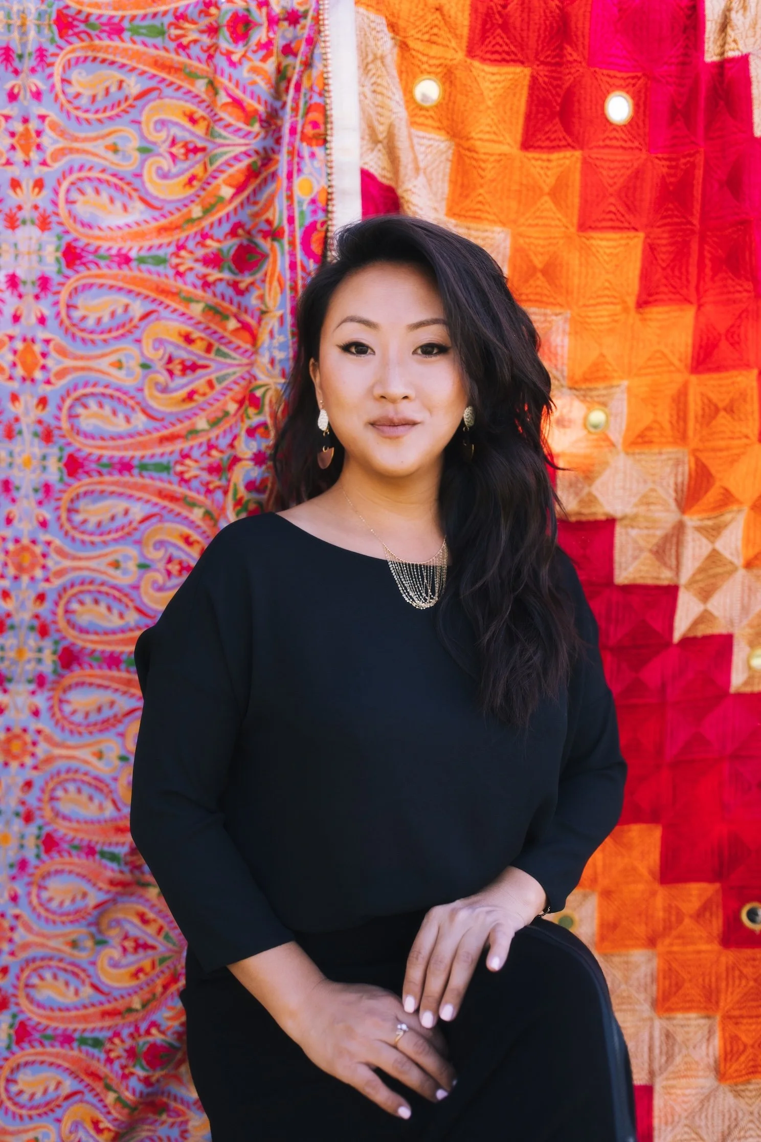A woman with dark hair wearing a black top and elegant jewelry, sitting in front of a vibrant, colorful fabric backdrop.