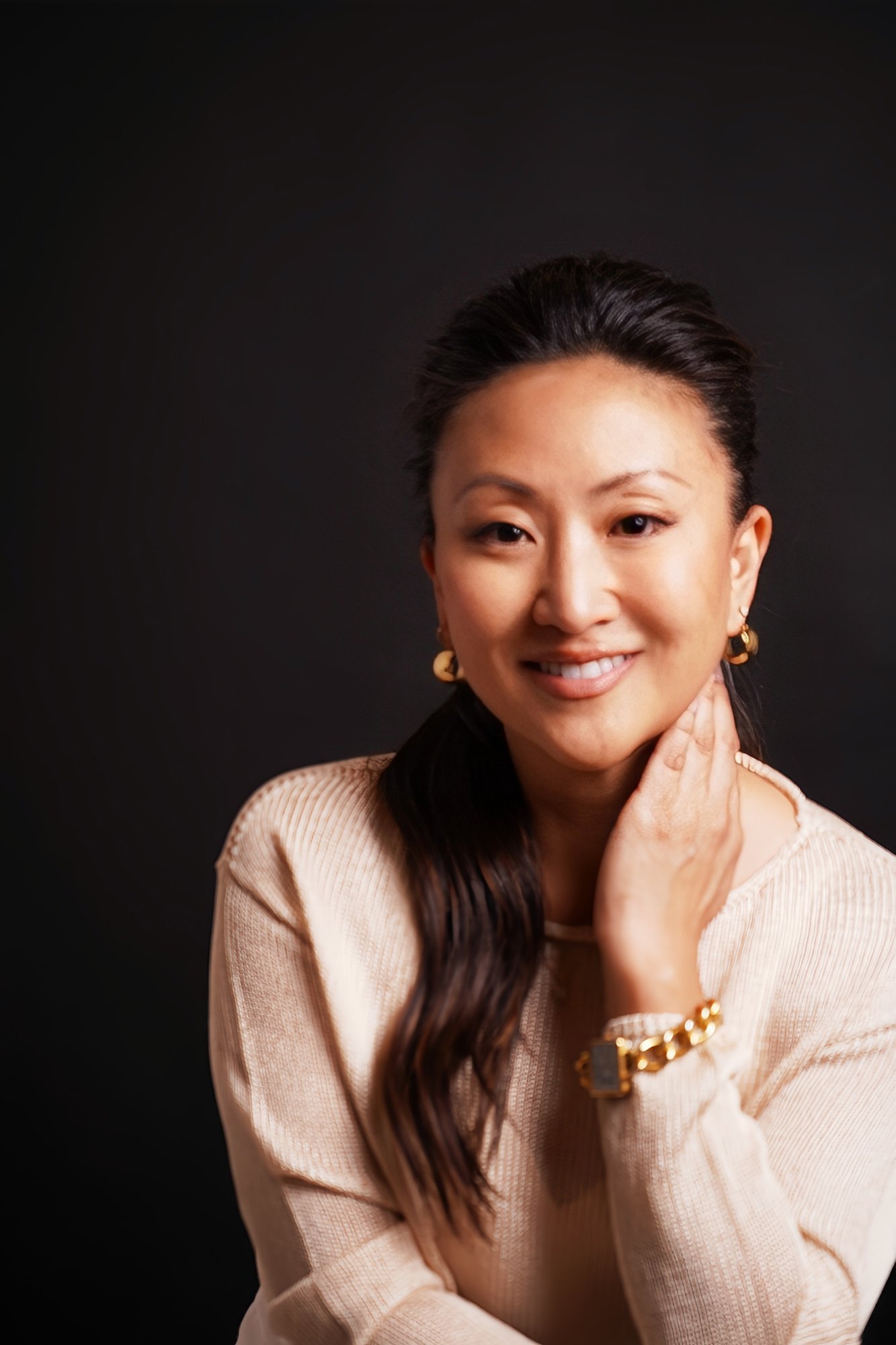 Portrait of a woman with long dark hair, wearing gold hoop earrings, a beige top, and a gold bracelet, smiling against a black background.