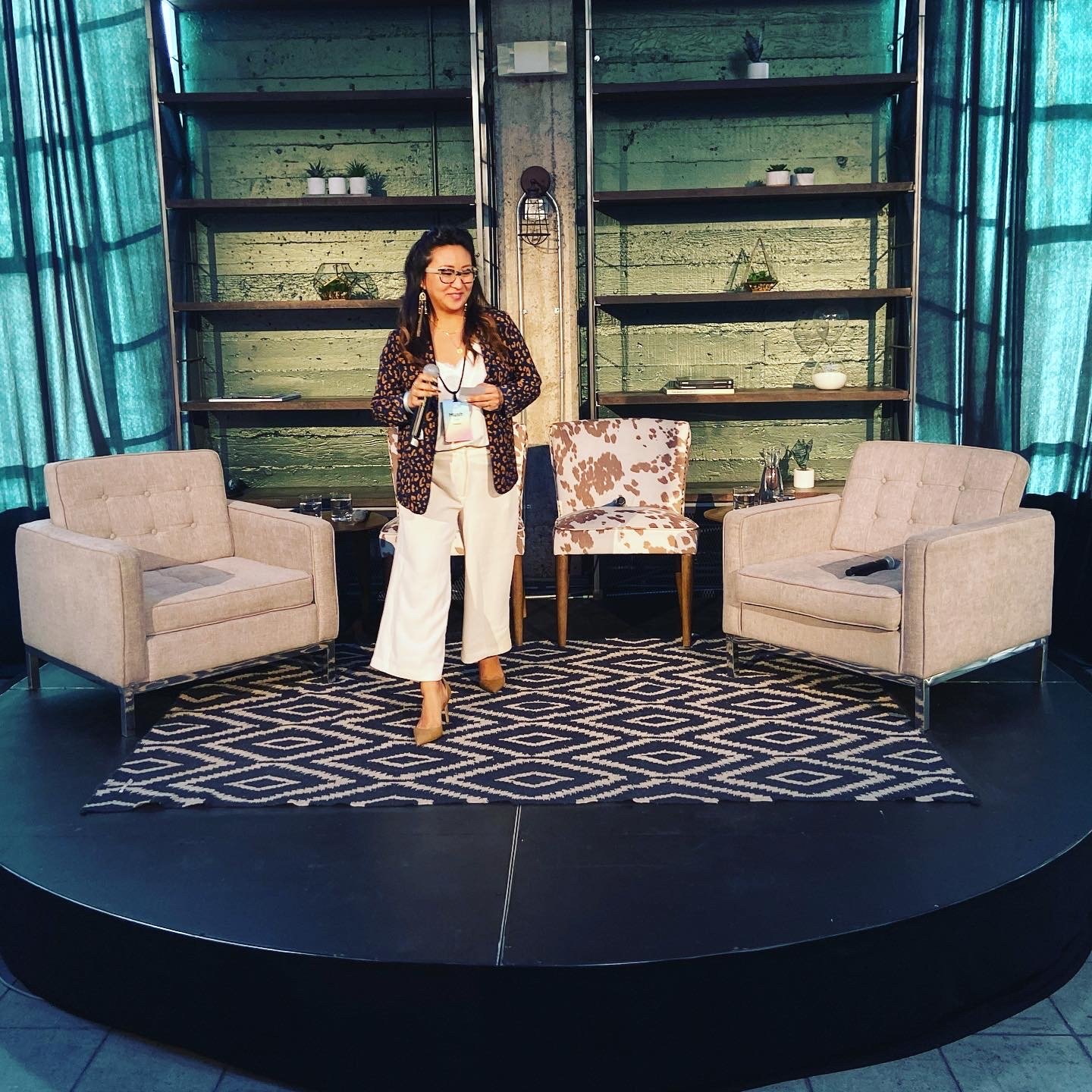 A woman standing on a stage with a microphone in her hand, in front of two beige armchairs and a patterned rug, with a green brick wall and shelves with plants and decorative items in the background.