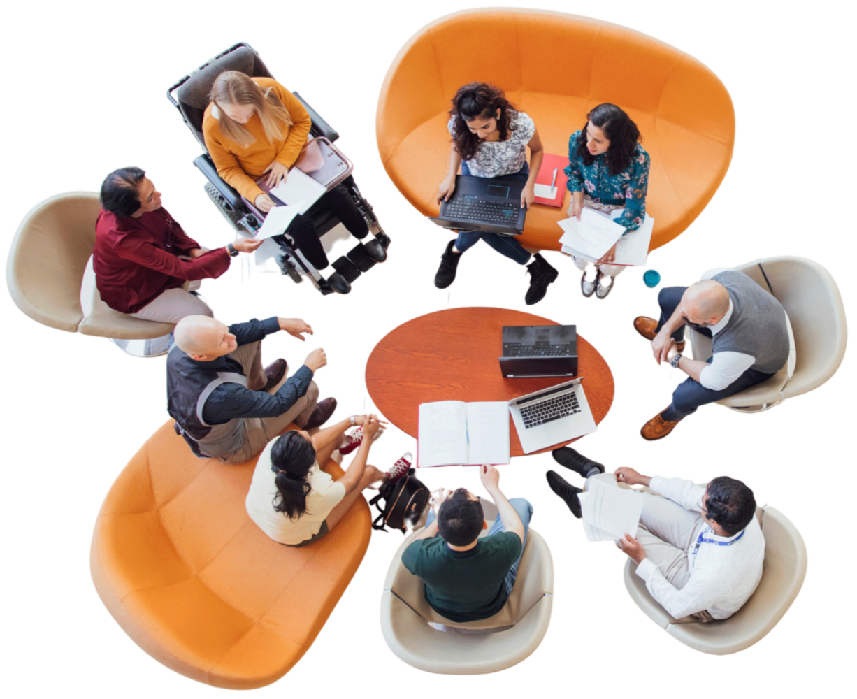 Overhead view of eight diverse people seated around a circular wooden table with laptops, notebooks, and papers, engaging in a meeting or discussion.