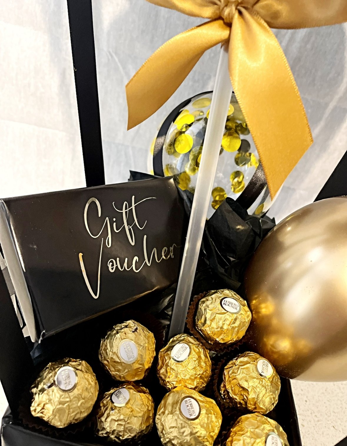 A gift basket containing gold-wrapped chocolates, a black gift voucher with white writing, a transparent ornament with yellow confetti, a large gold balloon, and a yellow ribbon.