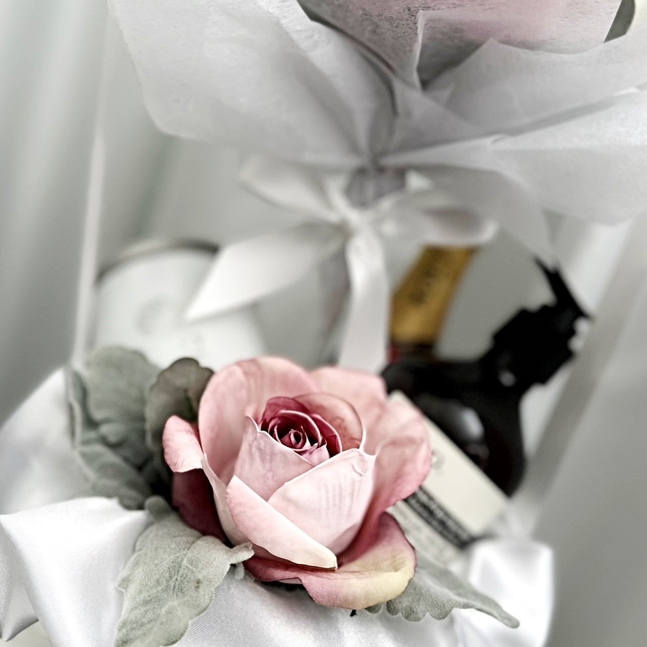 A pink and cream-colored artificial rose with green leaves resting on white satin fabric, with grocery items and a black object in the background.
