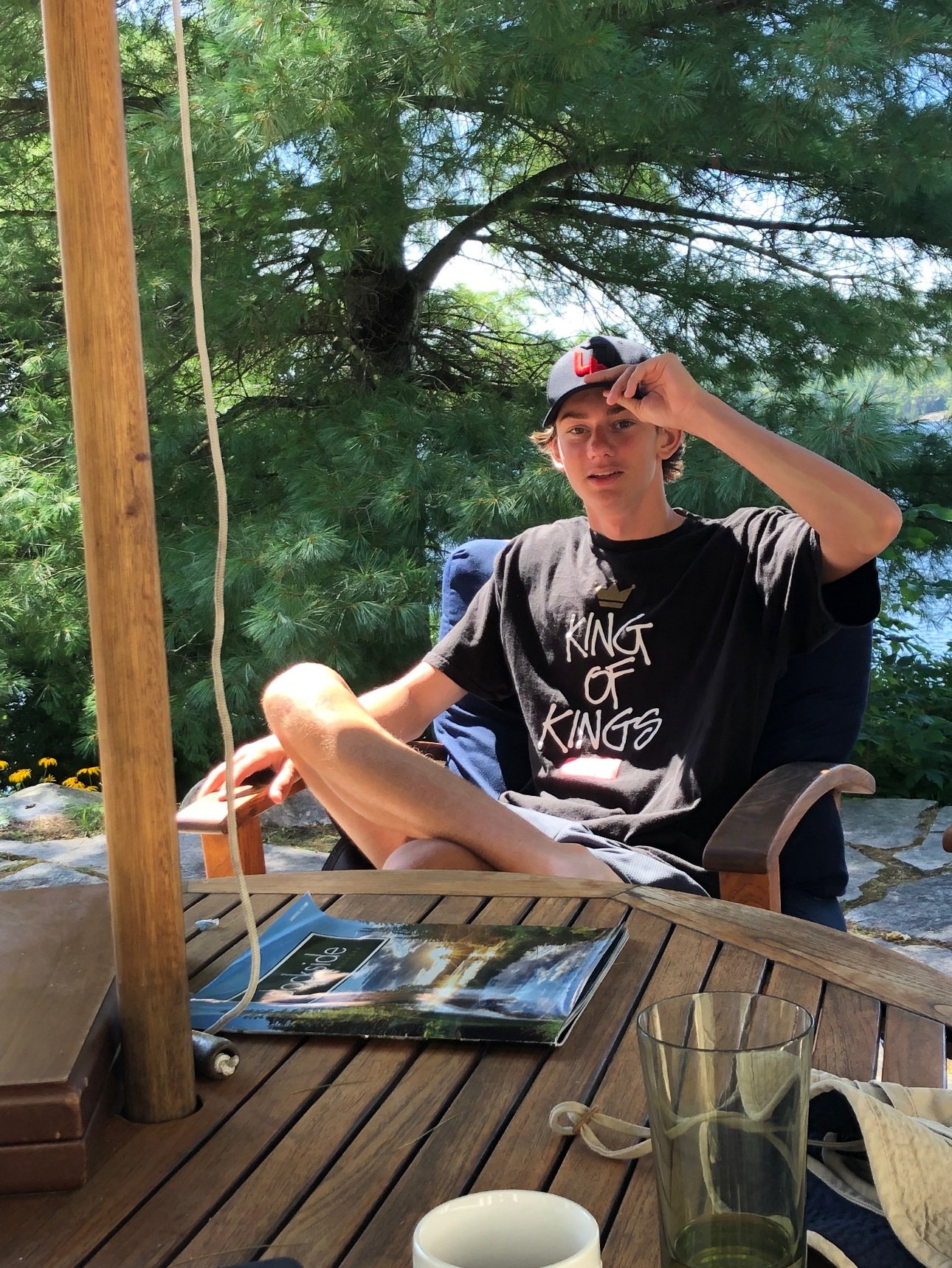 A young man sitting outdoors at a wooden table under a patio umbrella, wearing a black t-shirt with the words 'King of Kings' and a black cap with a red 'L' on it, holding the brim of his cap with one hand, surrounded by green foliage.