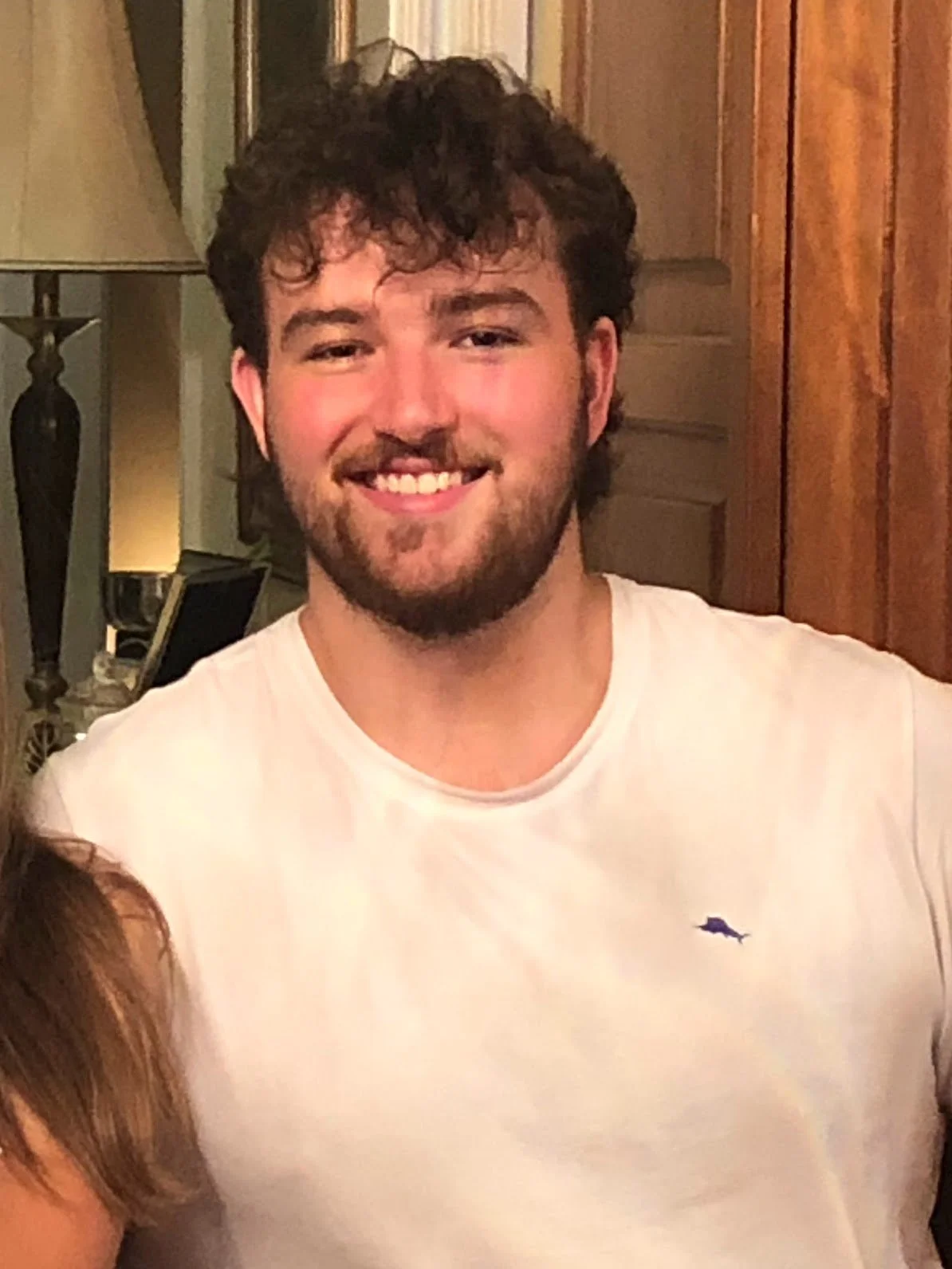 A young man with curly dark hair and a beard smiling, wearing a white shirt, in a warmly lit room with wooden furniture and a lamp.