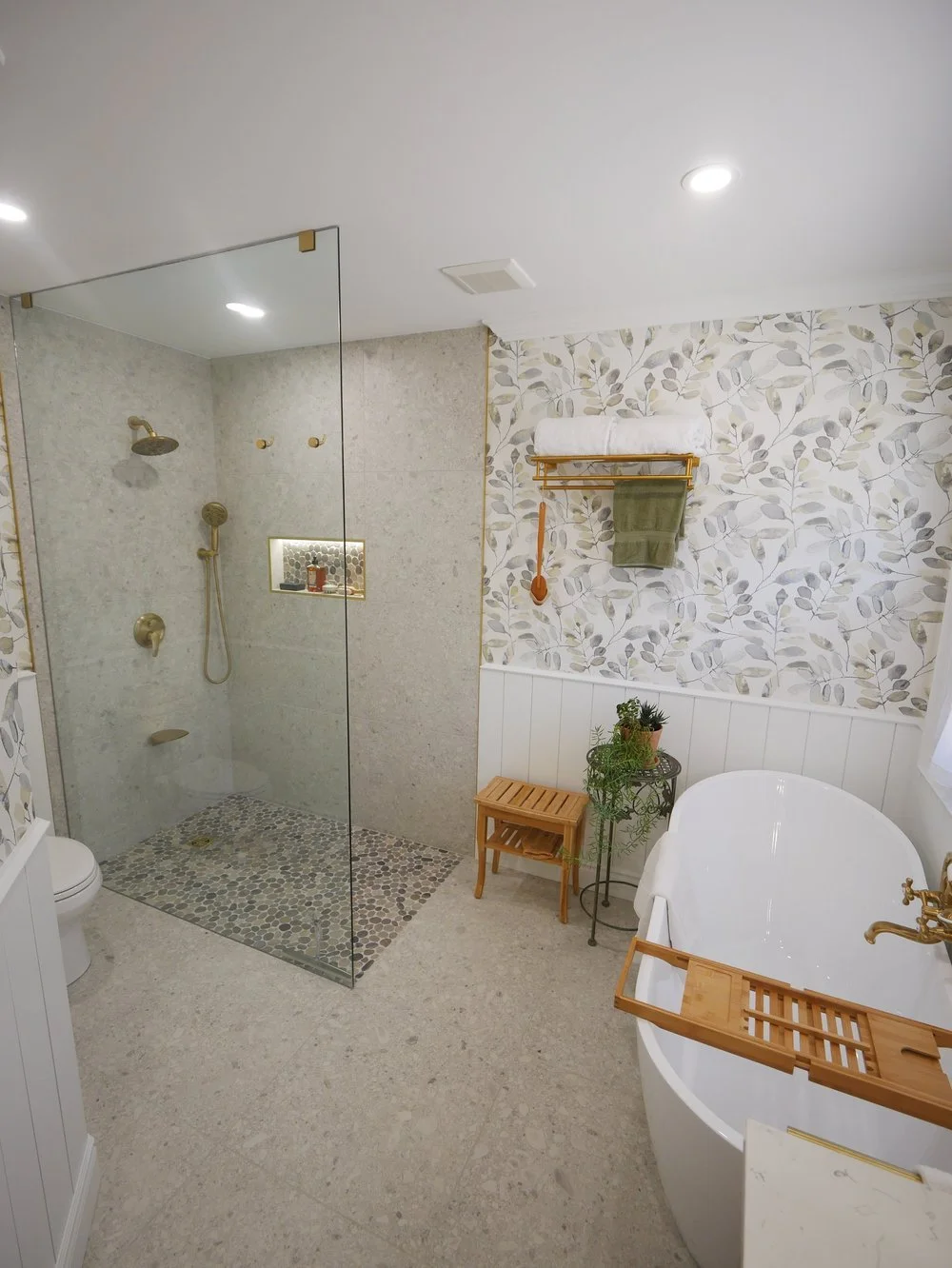 Bathroom with a walk-in shower, a white bathtub with a wooden tray, a small wooden stool, leafy wallpaper, green towels hanging, plants, and brass fixtures.