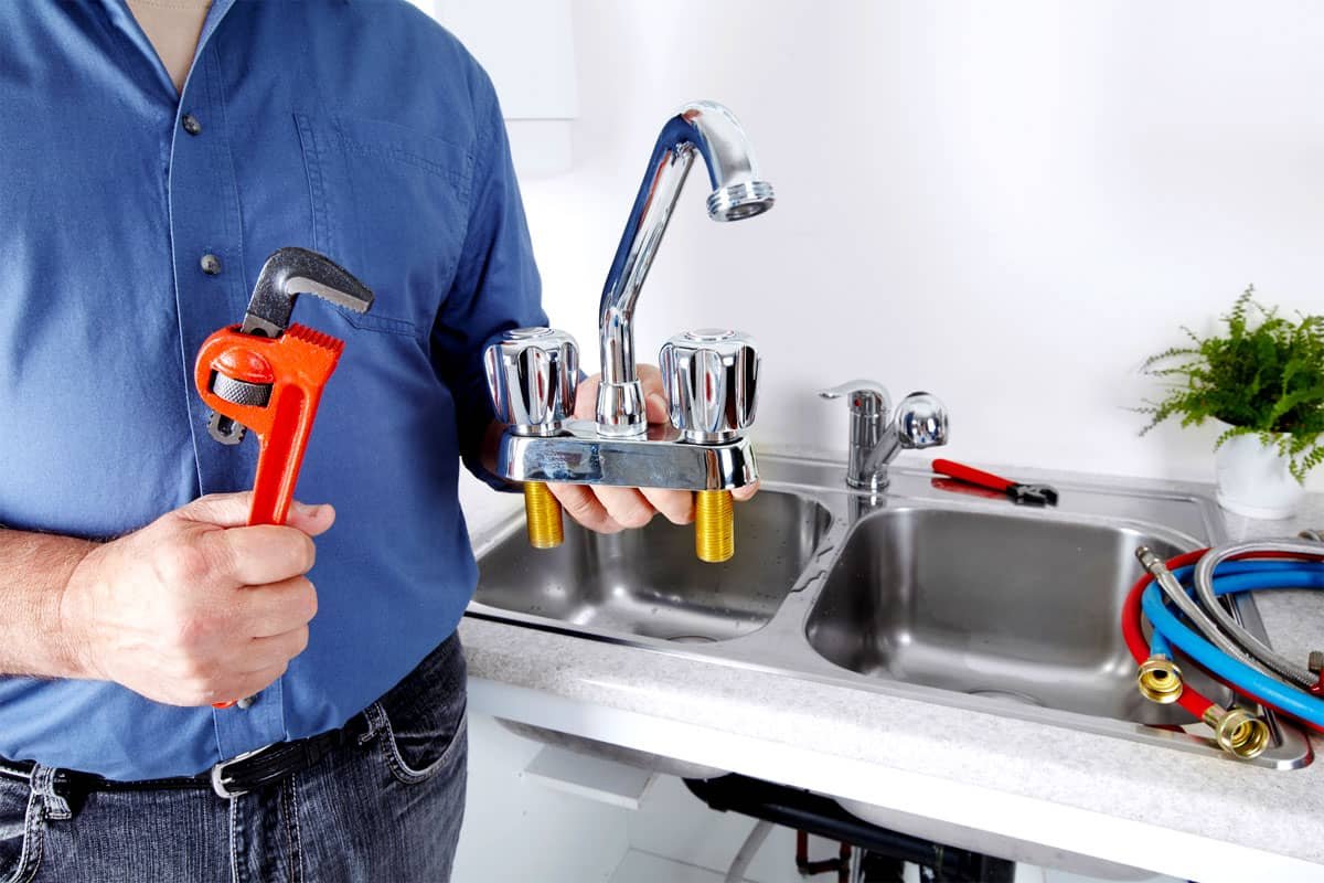 A person wearing a blue shirt holding a pipe wrench in one hand and a faucet fixture in the other, standing in front of a kitchen sink with plumbing hoses nearby.