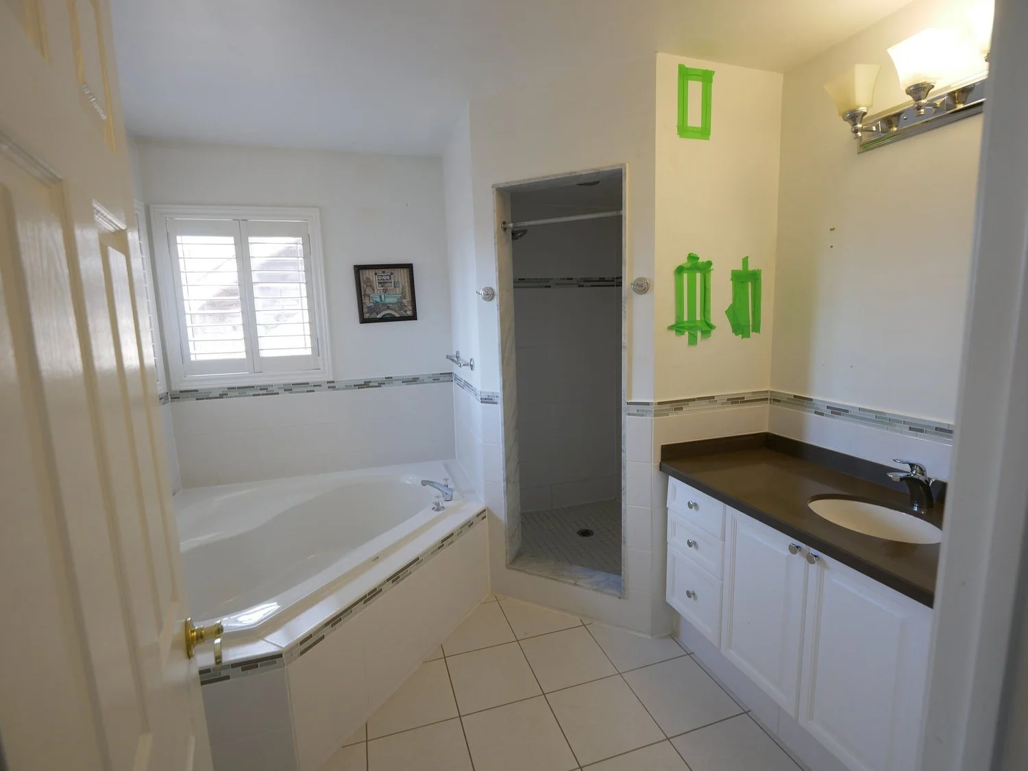 A bathroom with a bathtub, a window with shutters, a shower stall, a sink with a dark countertop, and marked areas on the wall for mirror and other fixtures, with mostly white walls and tiles.