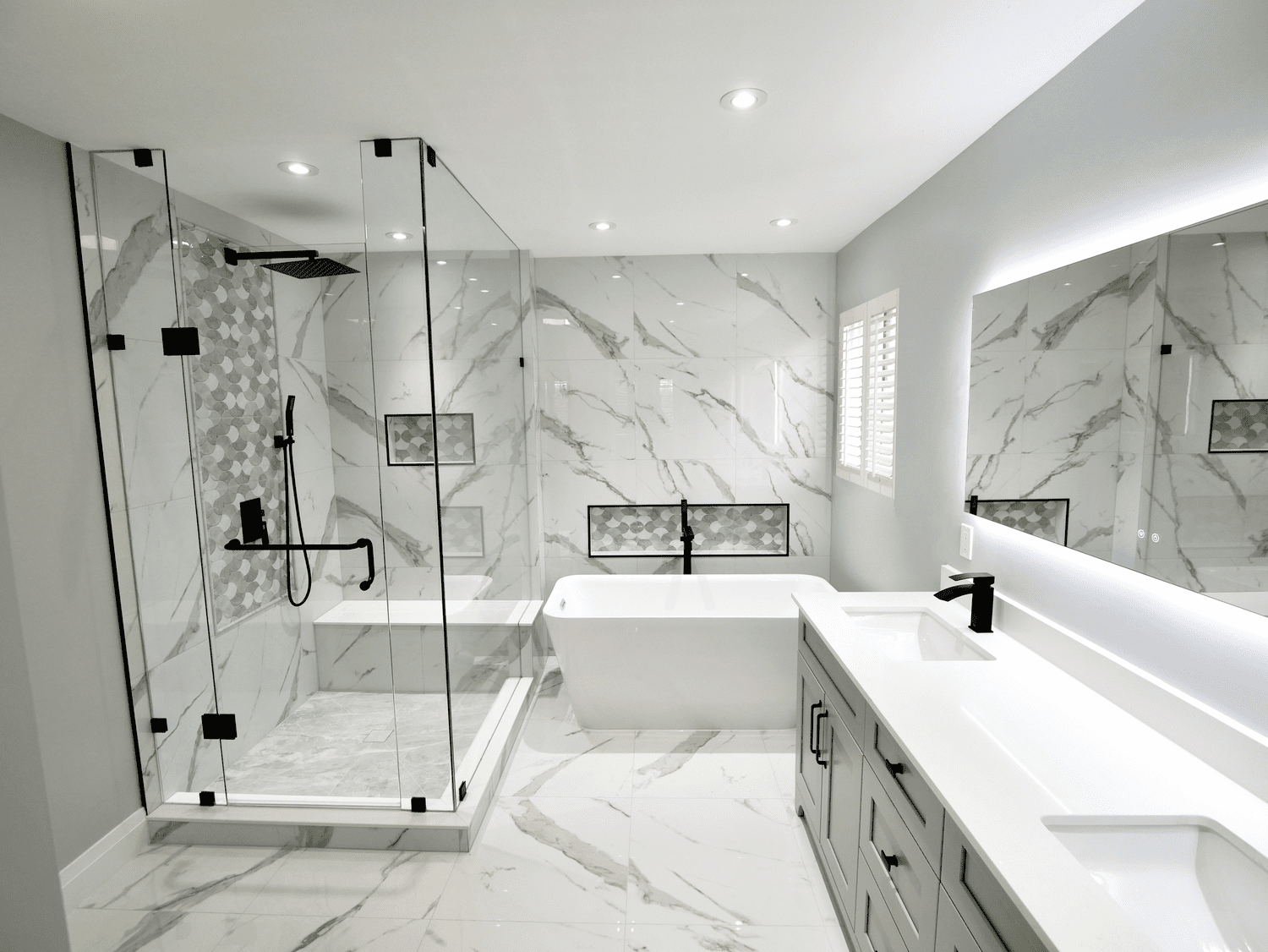 Modern bathroom with marble tiles, a glass shower enclosure, a bathtub, a double vanity with gray cabinets, a large mirror with LED lighting, and a window with shutters.