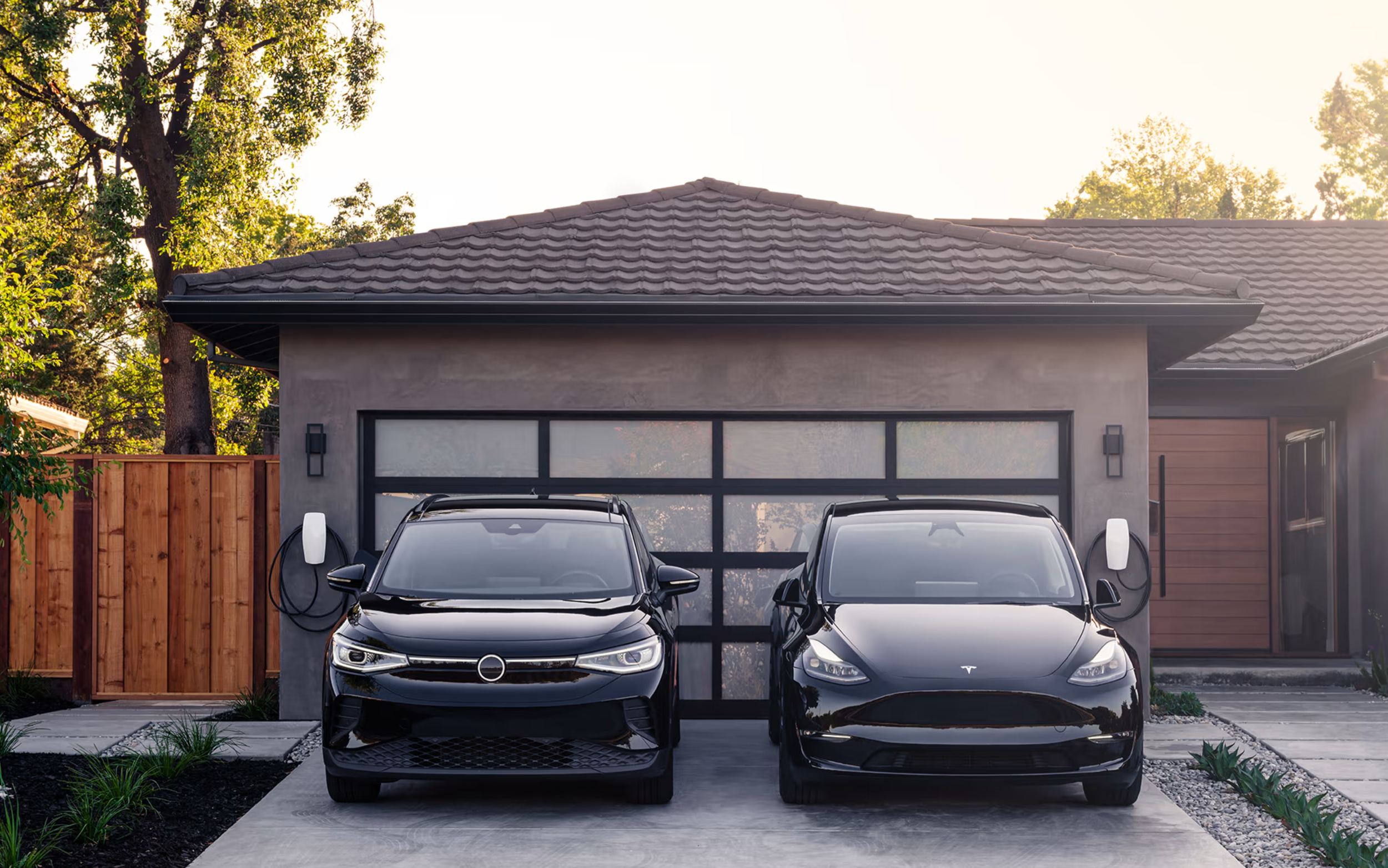 Modern house with two Tesla cars parked in driveway, solar panels on roof, surrounded by trees and a wooden fence.