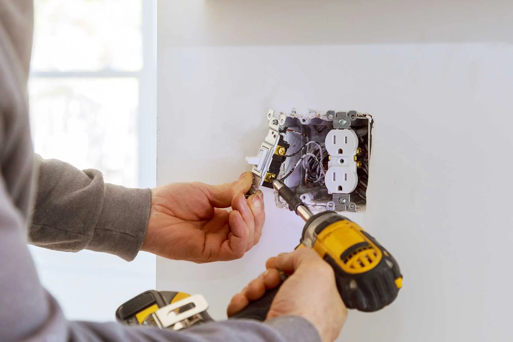 Person installing or repairing an electrical outlet using a cordless drill on a white wall.