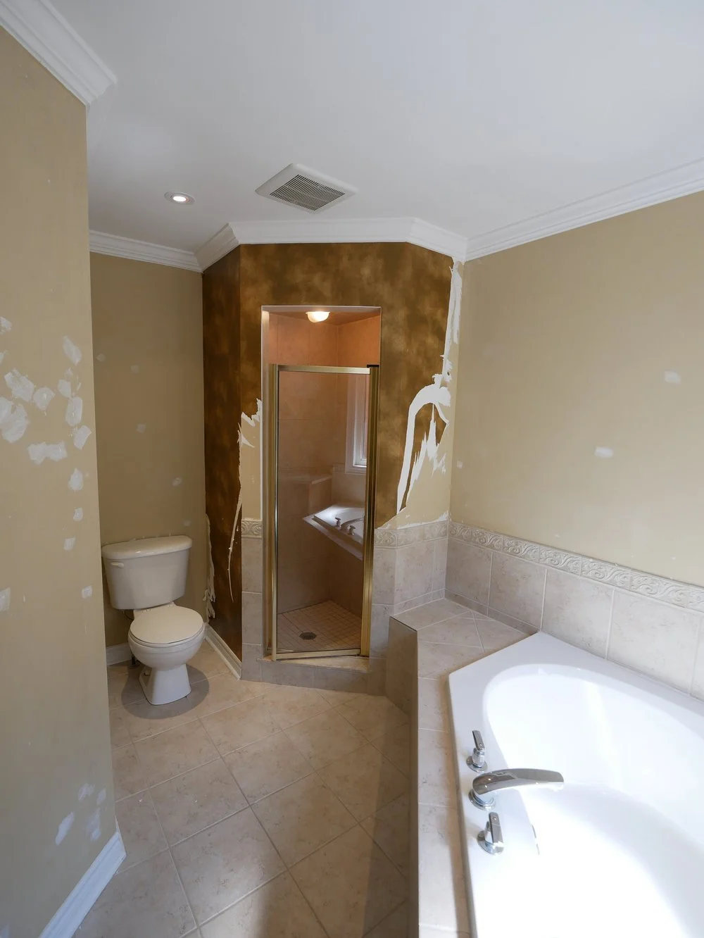 Bathroom with corner shower, toilet, built-in bathtub, and beige tile flooring and walls, during renovation.
