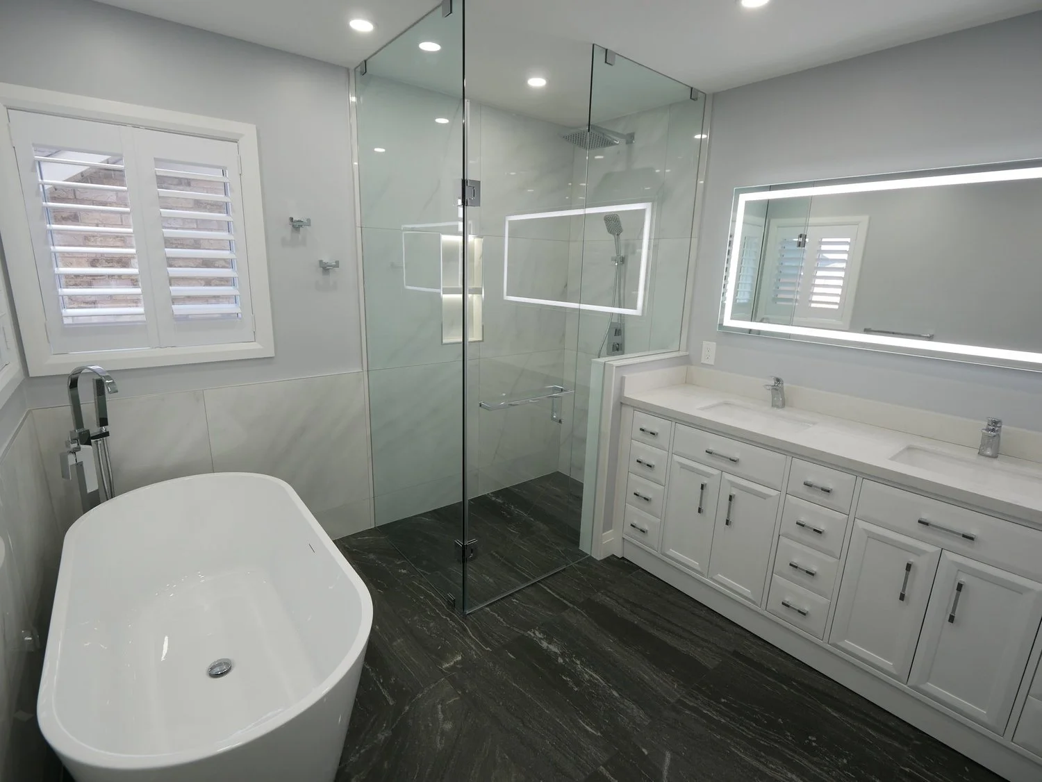 A modern bathroom with a white bathtub, a glass-enclosed walk-in shower with a rainfall showerhead, a large backlit mirror above a white vanity with double sinks, a window with blinds, and dark wood flooring.