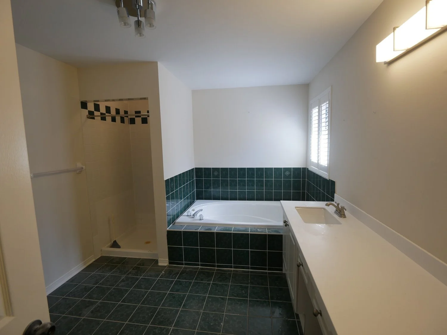 Bathroom with a bathtub, a small shower, a vanity with sink, green tiled walls and floors, and a window with shutters.