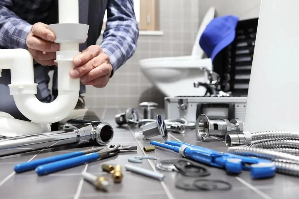 A person working on plumbing repairs with various plumbing tools and parts laid out on a work surface in a bathroom.