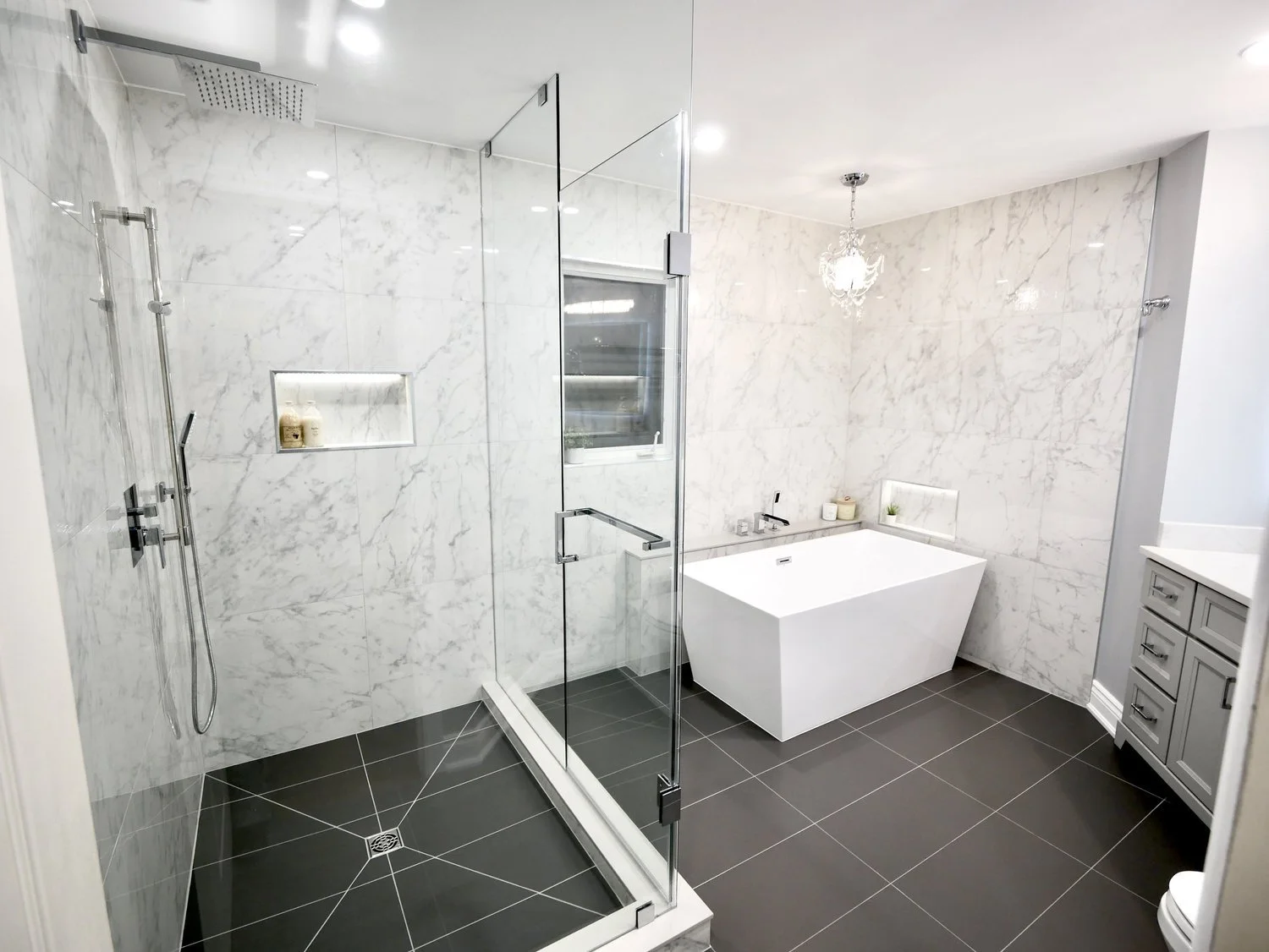 Modern bathroom with a walk-in shower with a glass door, marble tiles, a freestanding bathtub, gray cabinets, and a small window with a chandelier overhead.