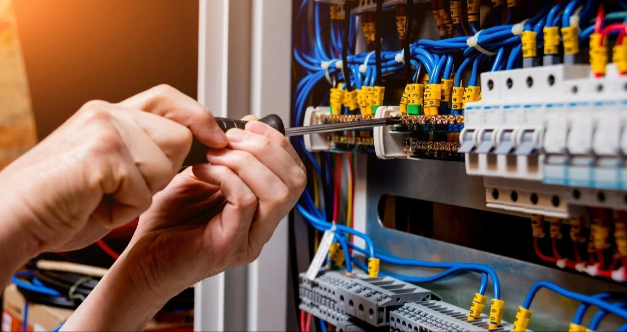 Close-up of a person's hands working on an electrical panel using a screwdriver, with numerous blue wires and yellow wire connectors inside the circuit box.